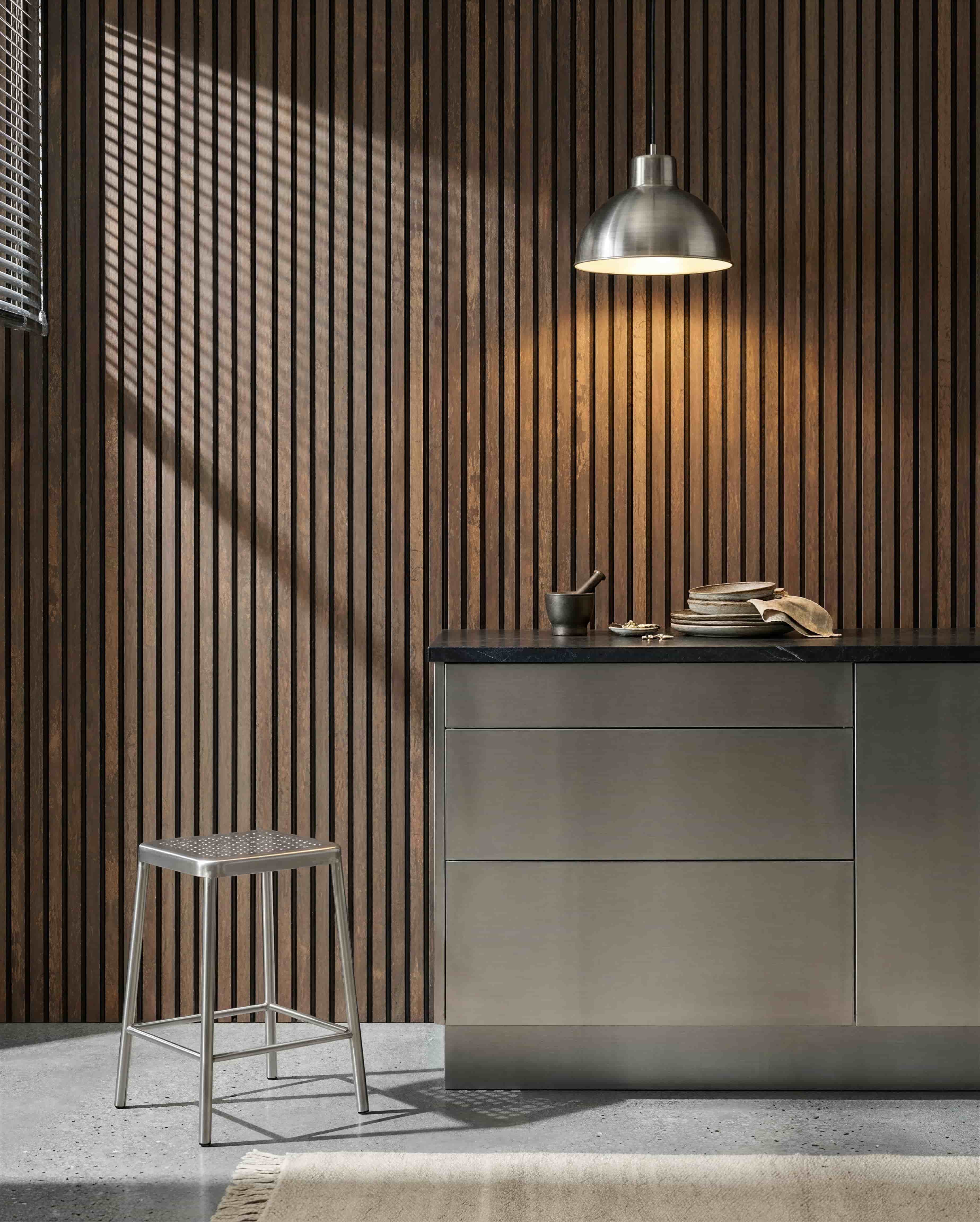 Küche mit einer WoodUpp Akupanel Oxide Copper Akustik-Lamellenwand, Arbeitsplatte aus Edelstahl, Metallhocker, Pendelleuchte, verschiedenen Geschirrteilen sowie Mörser und Stößel.