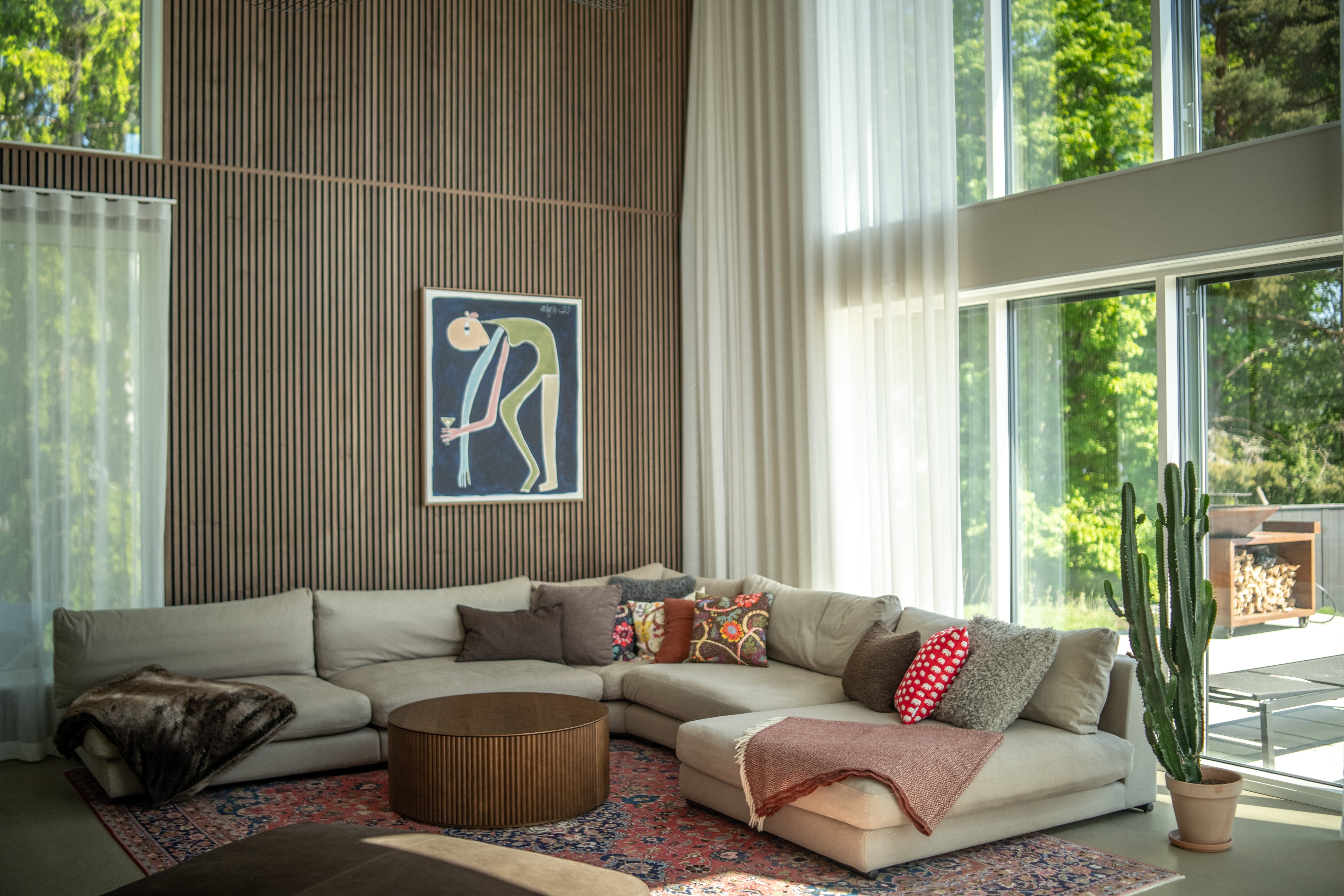 Modernes Wohnzimmer mit einem großen beigen Sektionssofa, bunten Kissen, rundem Couchtisch, Kaktus und raumhohen Fenstern mit transparenten Vorhängen und nordischen Akustikpaneelen.