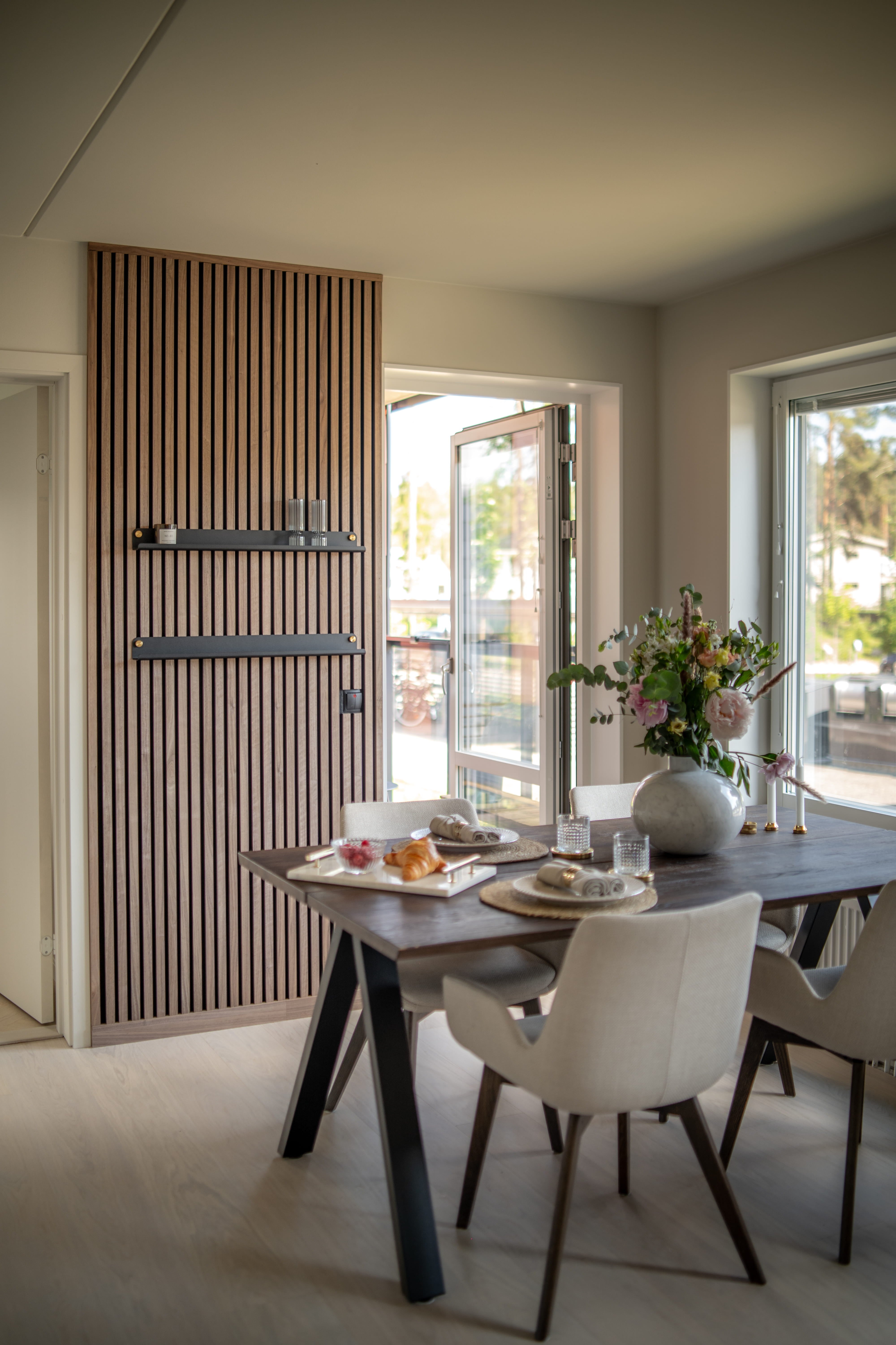 Modernes Esszimmer mit strukturierter Akupanel-Akustikholz-Lattenwand, dunklem Frühstückstisch, minimalistischen beigen Stühlen und einer Blumenvase vor großen offenen Fenstern.