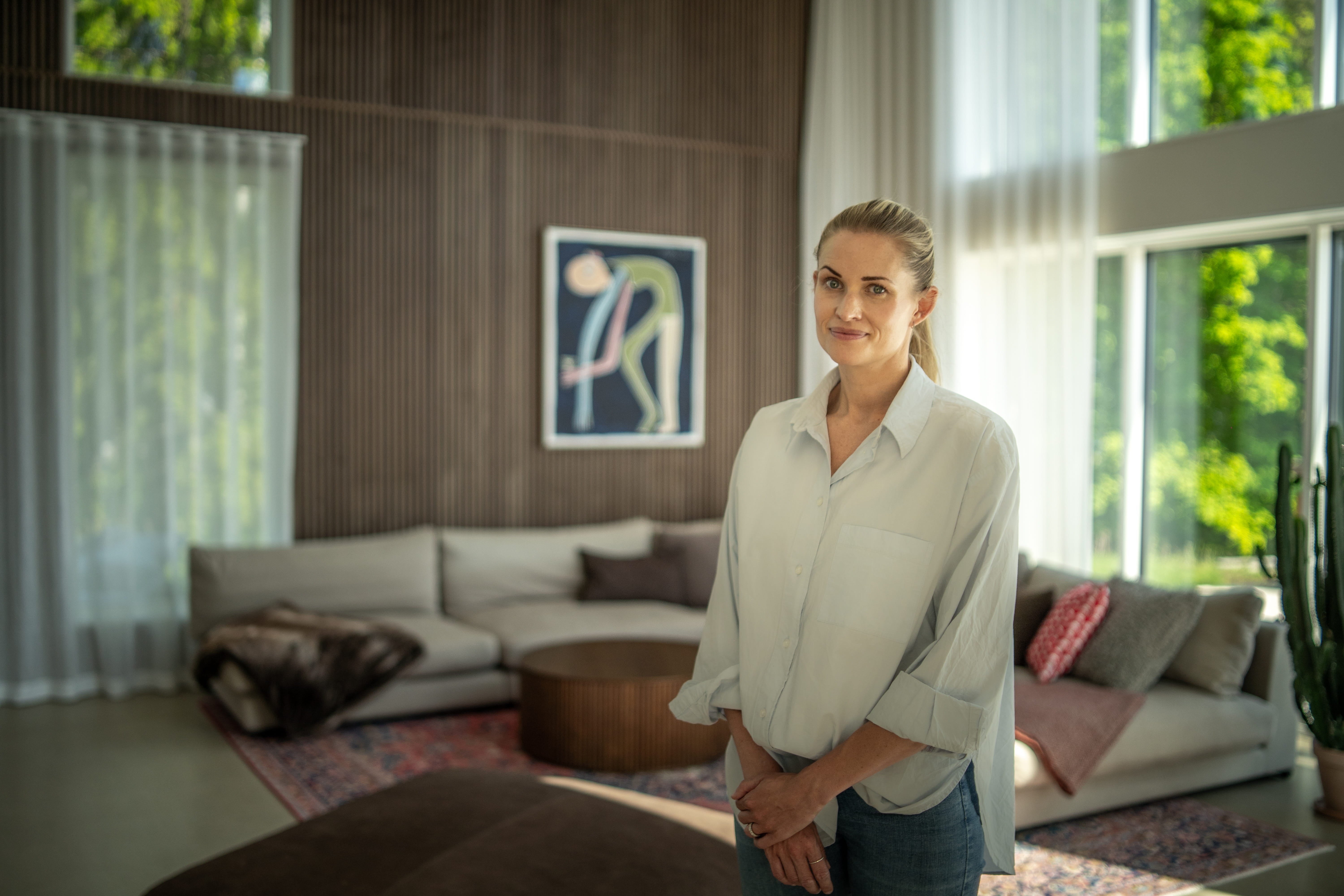 Eine Frau steht in einem sonnendurchfluteten Wohnzimmer mit großen Fenstern, nordischen Akustikholzpaneelen an der Wand, zeitgenössischer Kunst und minimalistischen Möbeln in neutralen Farbtönen.
