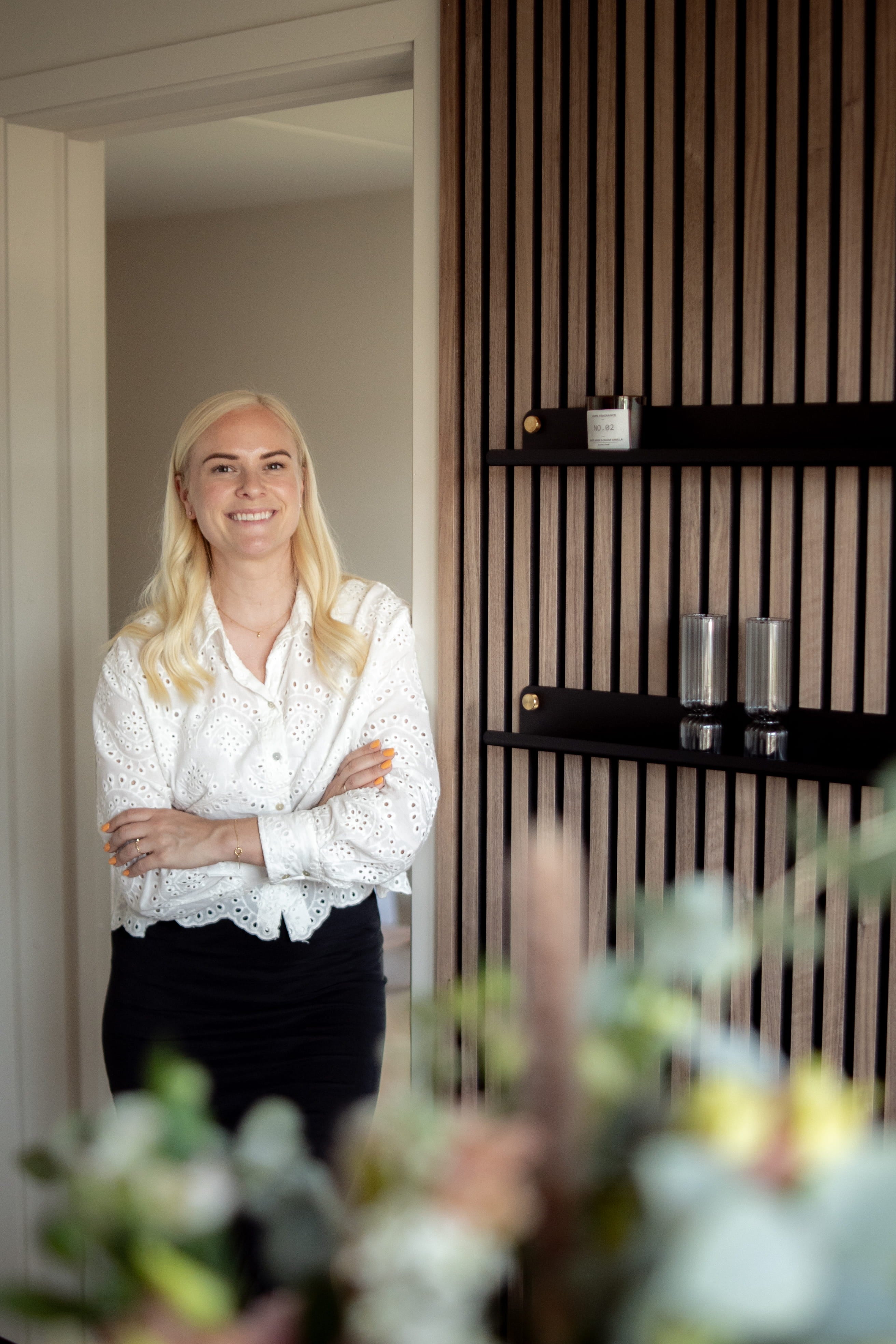 Eine Frau mit langen blonden Haaren und weißer Bluse steht lächelnd mit verschränkten Armen in einem modernen Raum mit nordischen Akustikholzpaneelen und schlichten Regalen.