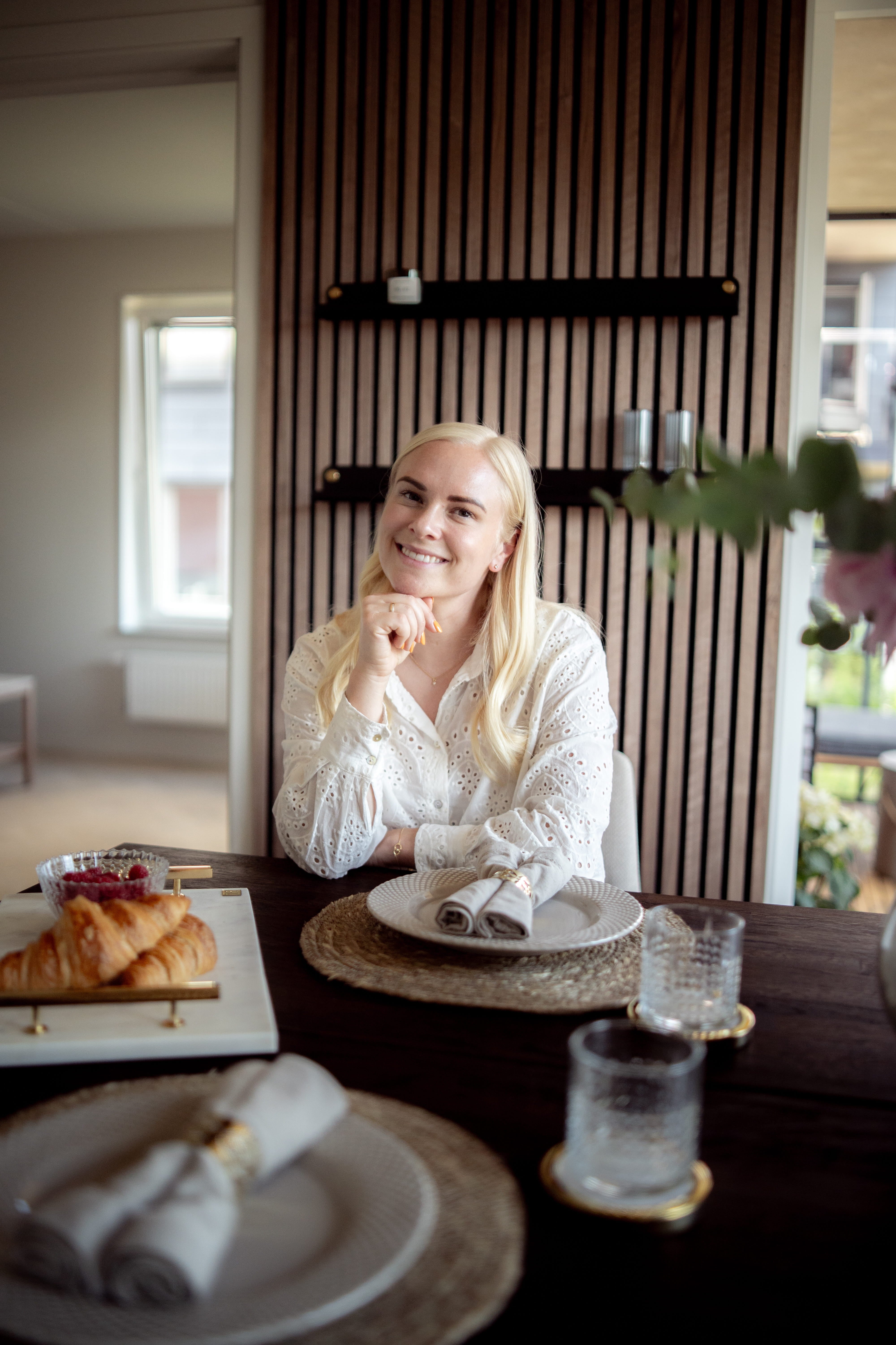 Eine Frau mit langen blonden Haaren sitzt an einem für das Frühstück gedeckten Esstisch mit Croissants und Gläsern, neben einer nordischen Akustikplatte an der Wand.
