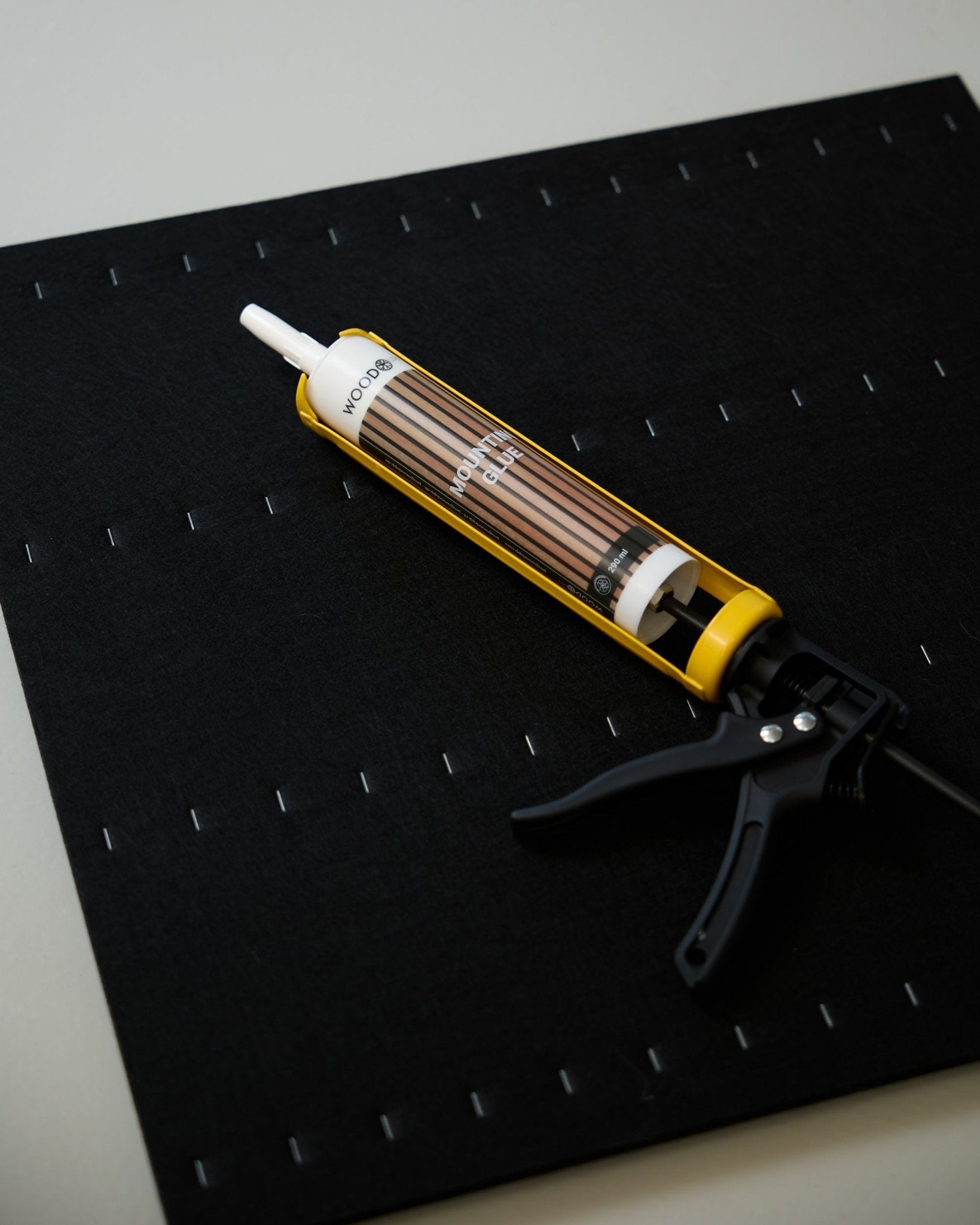 Eine gelbe und black WoodUpp Kartuschenpistole (290 ml) liegt auf einer black Matte mit einem Raster aus weißen Klammern und wird für die Montage von Akustikpaneelen verwendet.