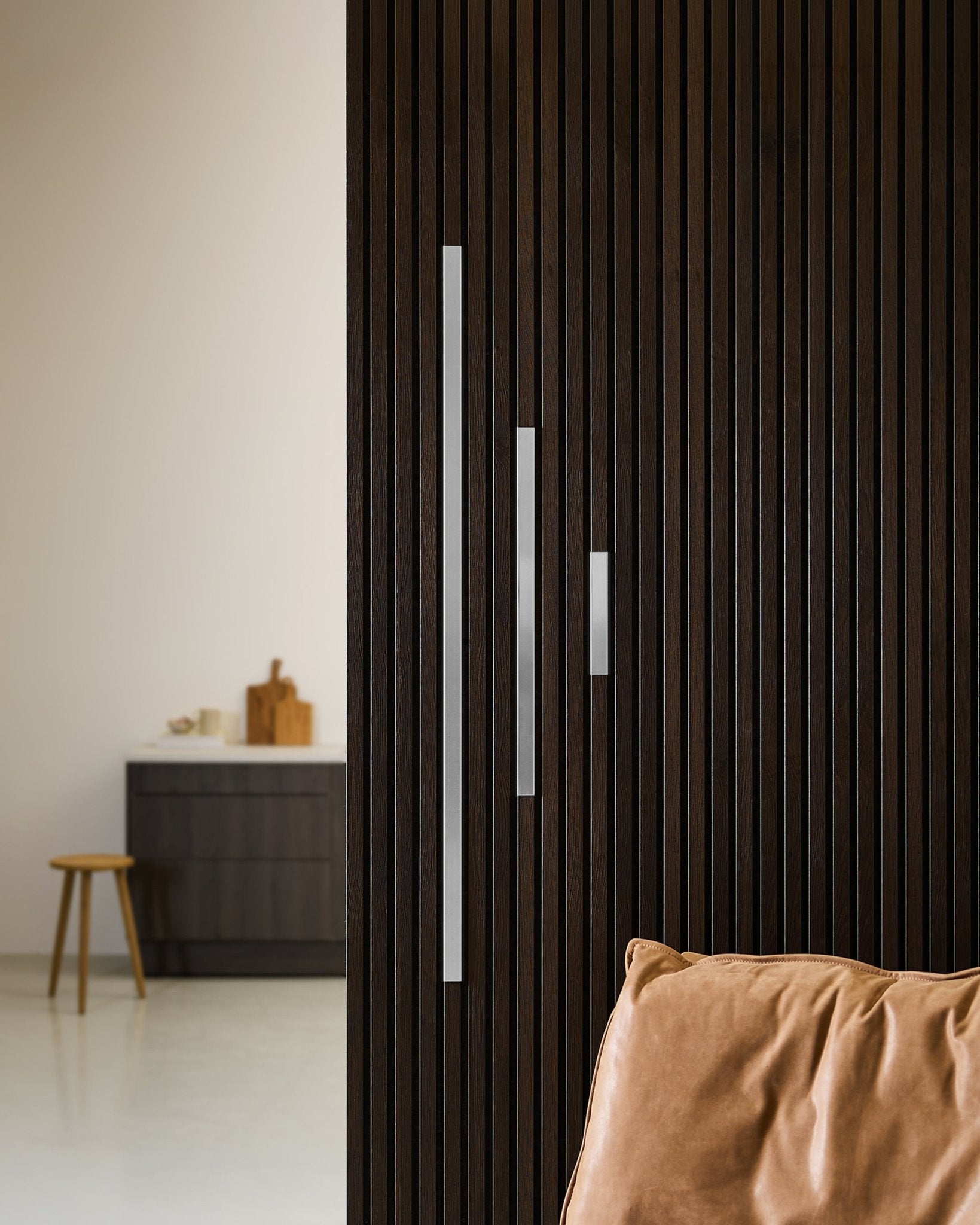 Minimalistisches Interieur mit einer Akustikpaneel Wand aus dunklem Holz, einer WoodUpp Liva Cover in poliertem steel (20 cm), einem hellbraunen Kissen und modernen Möbeln.