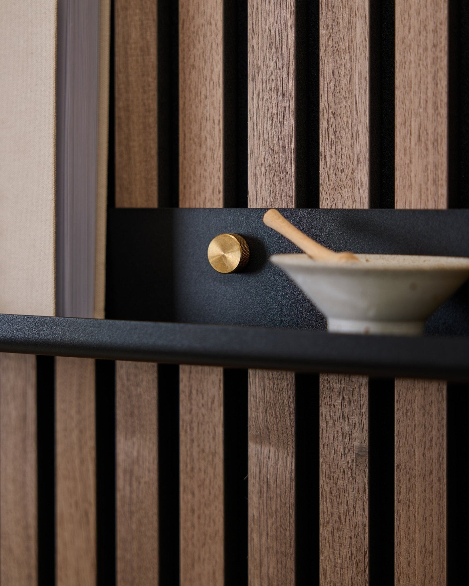Nahaufnahme eines 115,9 cm langen black schmalen Regals Norr von WoodUpp, montiert an einer nordischen Akustik-Lamellenwand mit brass Rändelschrauben, goldenem Knauf und Keramikschale mit Löffel.