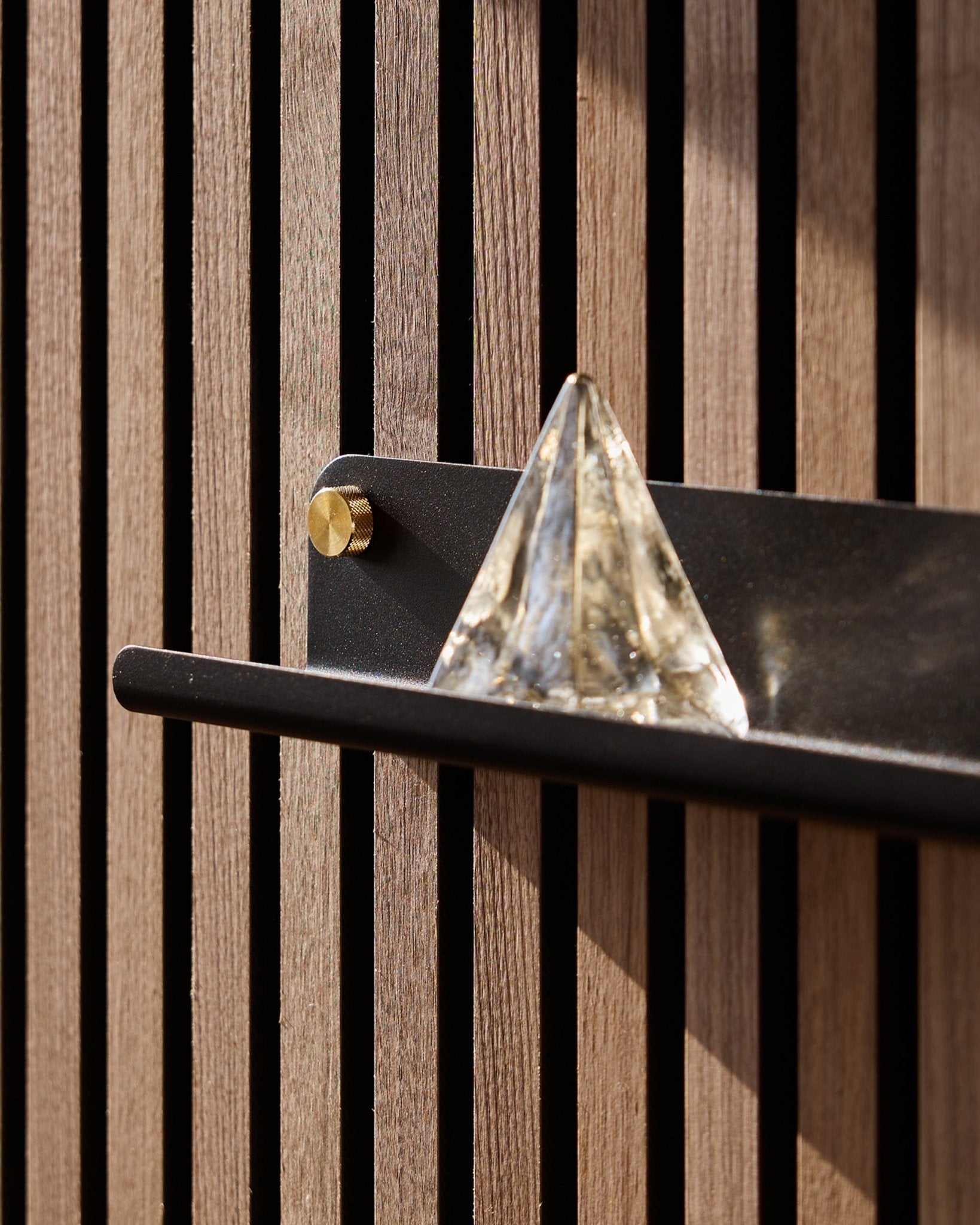 Eine Glaspyramide steht auf dem WoodUpp Norr Narrow Shelf in black (51,9 cm), montiert an einer vertical Akustikpaneel und vom Sonnenlicht hervorgehoben.