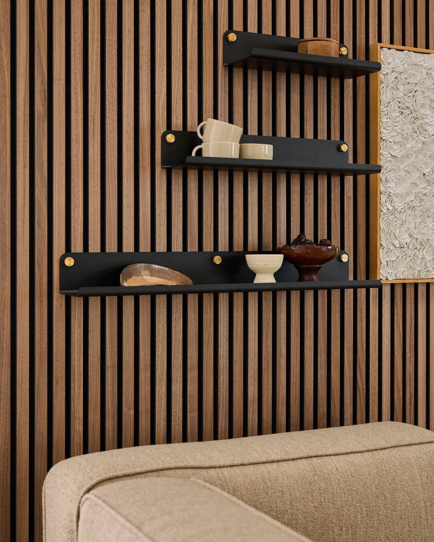 Drei black Regalböden, darunter das WoodUpp Norr Wide Shelf (75,9 cm), präsentieren Dekor auf einer Holzlamellen-Akustikpaneel Wand über einem beige Sofa; ein Teil des Kunstwerks ist sichtbar.