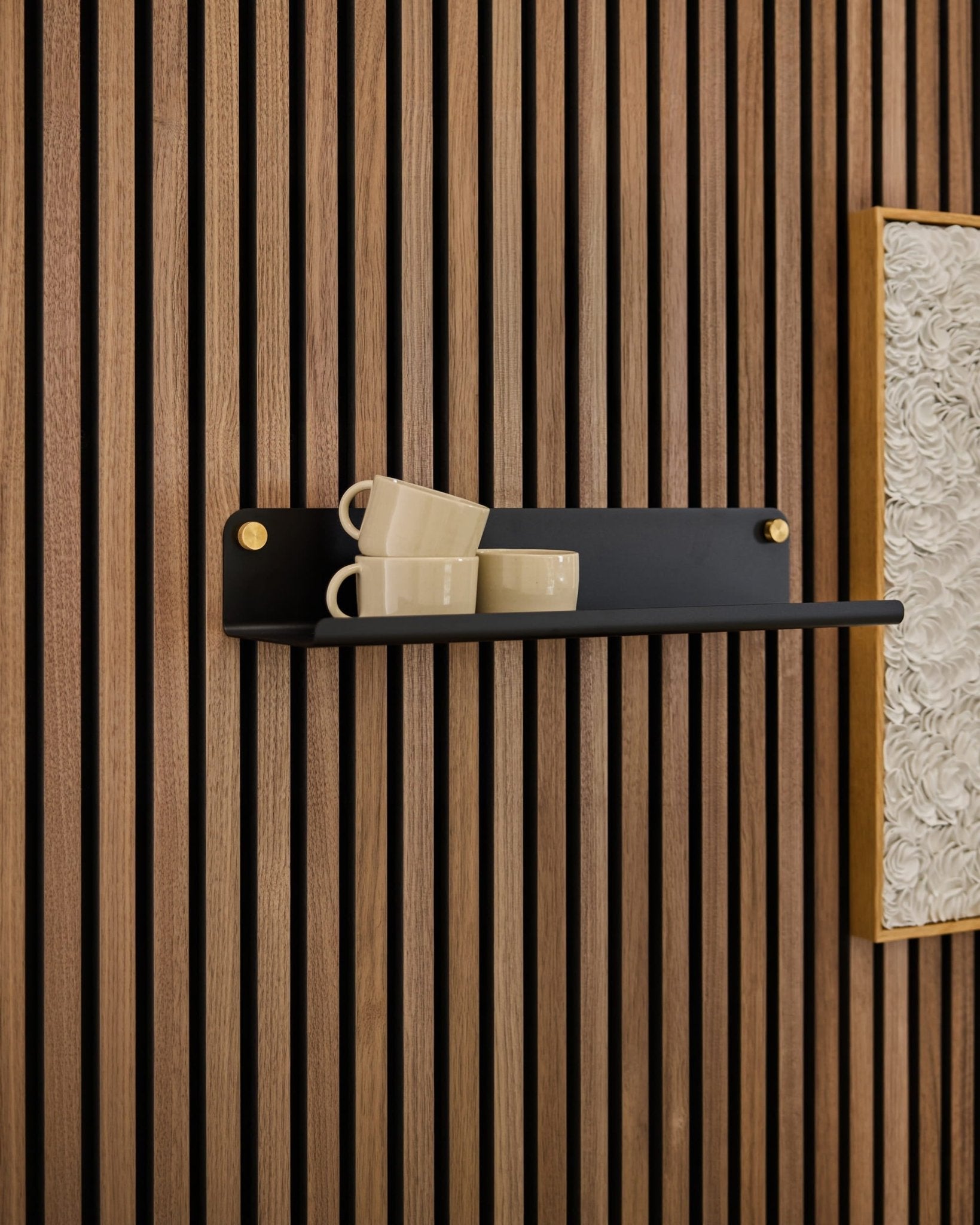 Auf einem schwarzen Norr Wide Shelf-Regal von WoodUpp stehen drei beigefarbene Tassen, das Regal ist an einer Akupanel-Akustikplatte neben einem gerahmten, strukturierten weißen Kunstwerk befestigt.
