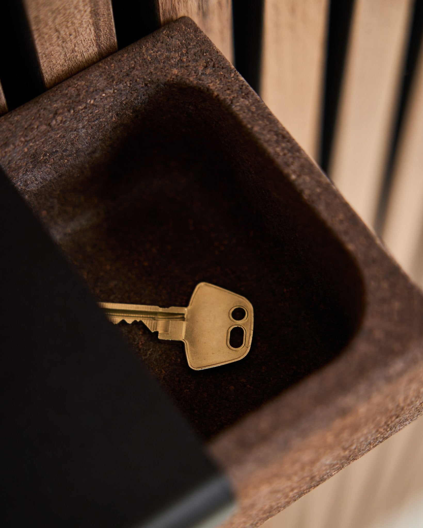 Ein brass Schlüssel liegt auf einem Norr Key Tray/Rauchkork, 35,9 cm von WoodUpp, platziert auf einem brown Behälter vor einem Hintergrund aus Akupanel Akustik-Holzpaneelen.