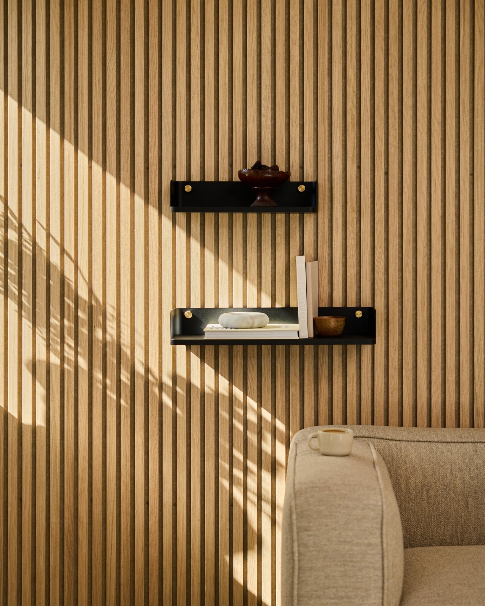 Sonnenlicht fällt auf ein beige Sofa und WoodUpps Linn Bücherregal in black oak, 55,9 cm, vor einer minimalistischen nordischen Lamellenwand Akustikpaneel.