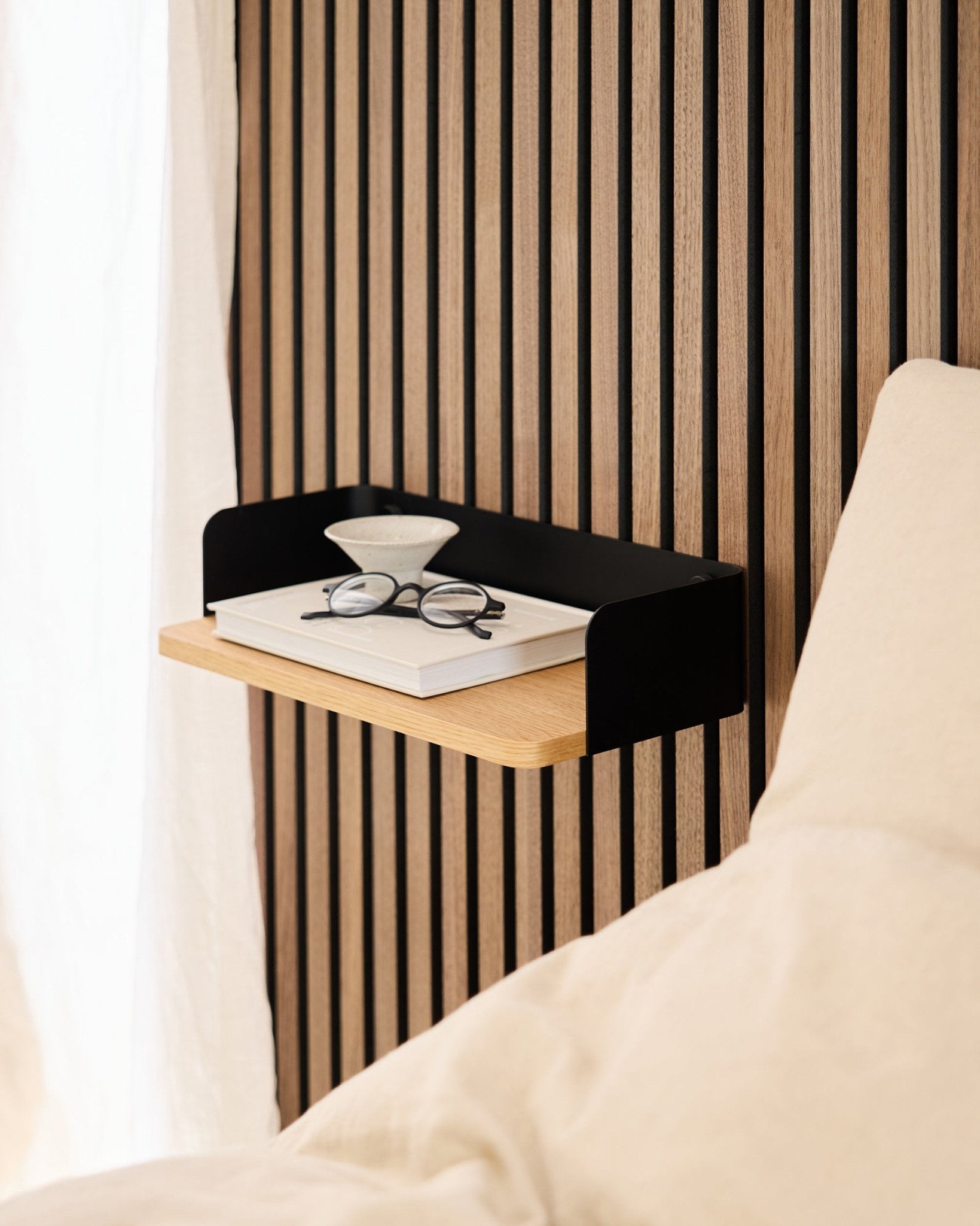 Das Linn Bücherregal von WoodUpp in natural oak (39,9 cm) mit einem Buch, einer Brille und einem Glas neben einem Bett vor einer Wand aus Akupanel Akustikholzpaneelen.