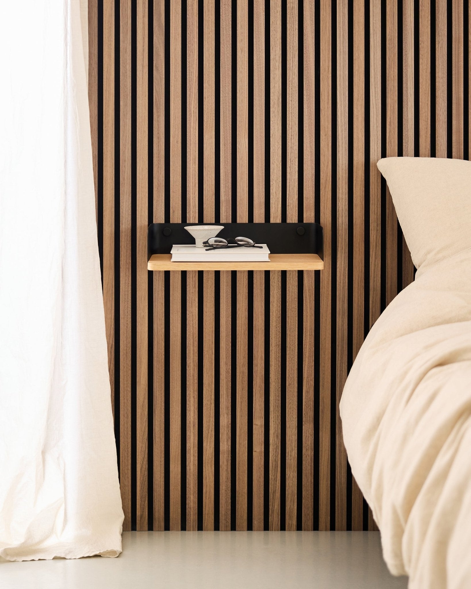 Minimalistisches Schlafzimmer mit einer nordischen Holzlamellenwand in Akustikpaneel, dem WoodUpp Linn Bücherregal in Natural Oak und einem schwebenden Nachttisch neben einem Bett in beige.