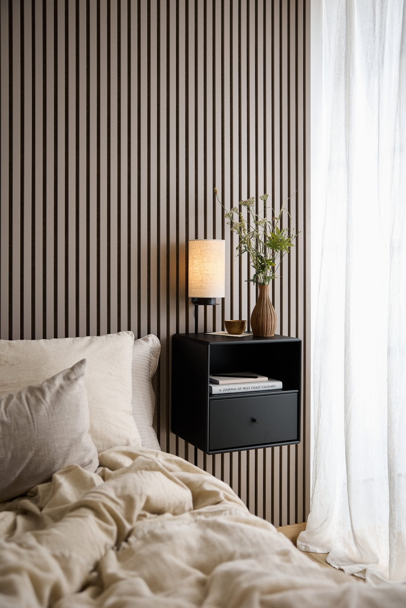 Ein Bett mit beiger Bettwäsche, ein schwarzes WoodUpp Arka-Bücherregal mit Schublade, eine Lampe und eine Vase neben einem Fenster mit weißen, durchsichtigen Vorhängen.