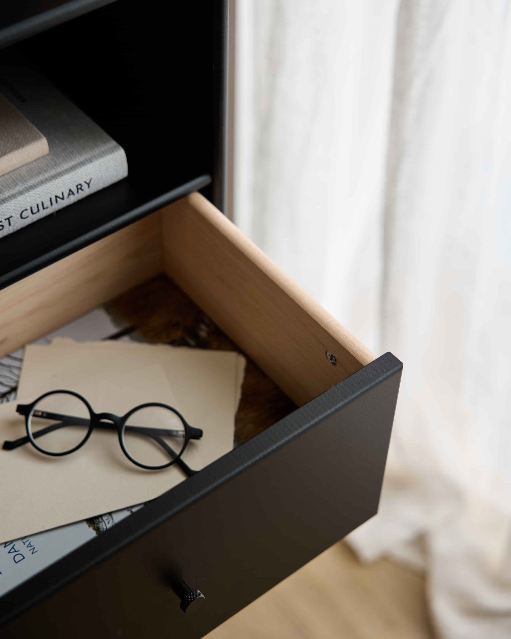 Eine runde schwarze Brille und Papiere in einer offenen WoodUpp Arka | Bücherregal-Schublade / Schwarz, unter Büchern, während transparente Vorhänge das Hintergrundlicht sanft streuen.