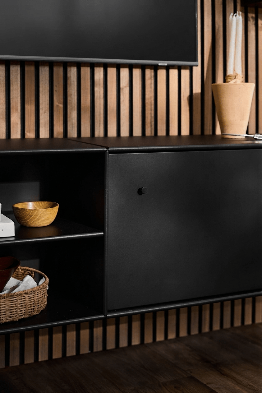 TV-Konsole „Black WoodUpp Arka“ mit offenen Regalen für Bücher und Körbe, hellbrauner Vase, praktischer Kabelführung und vertikaler Akupanel-Akustikwand aus Holz im Hintergrund.