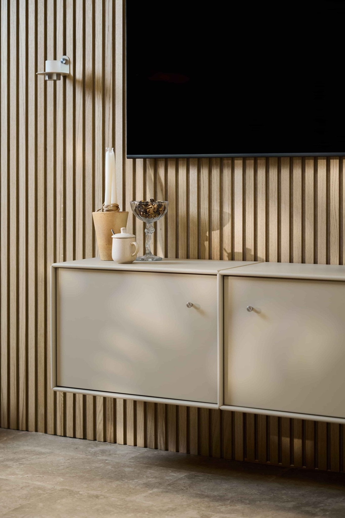 Minimalistisches Wohnzimmer mit dem TV-Schrank „Arka“ von WoodUpp in Beige, einer Akupanel-Akustikwand aus Holz, einem an der Wand montierten Fernseher, Kerzen, einer Glasschale, einem weißen Gefäß und ordentlich verlegten Kabeln.