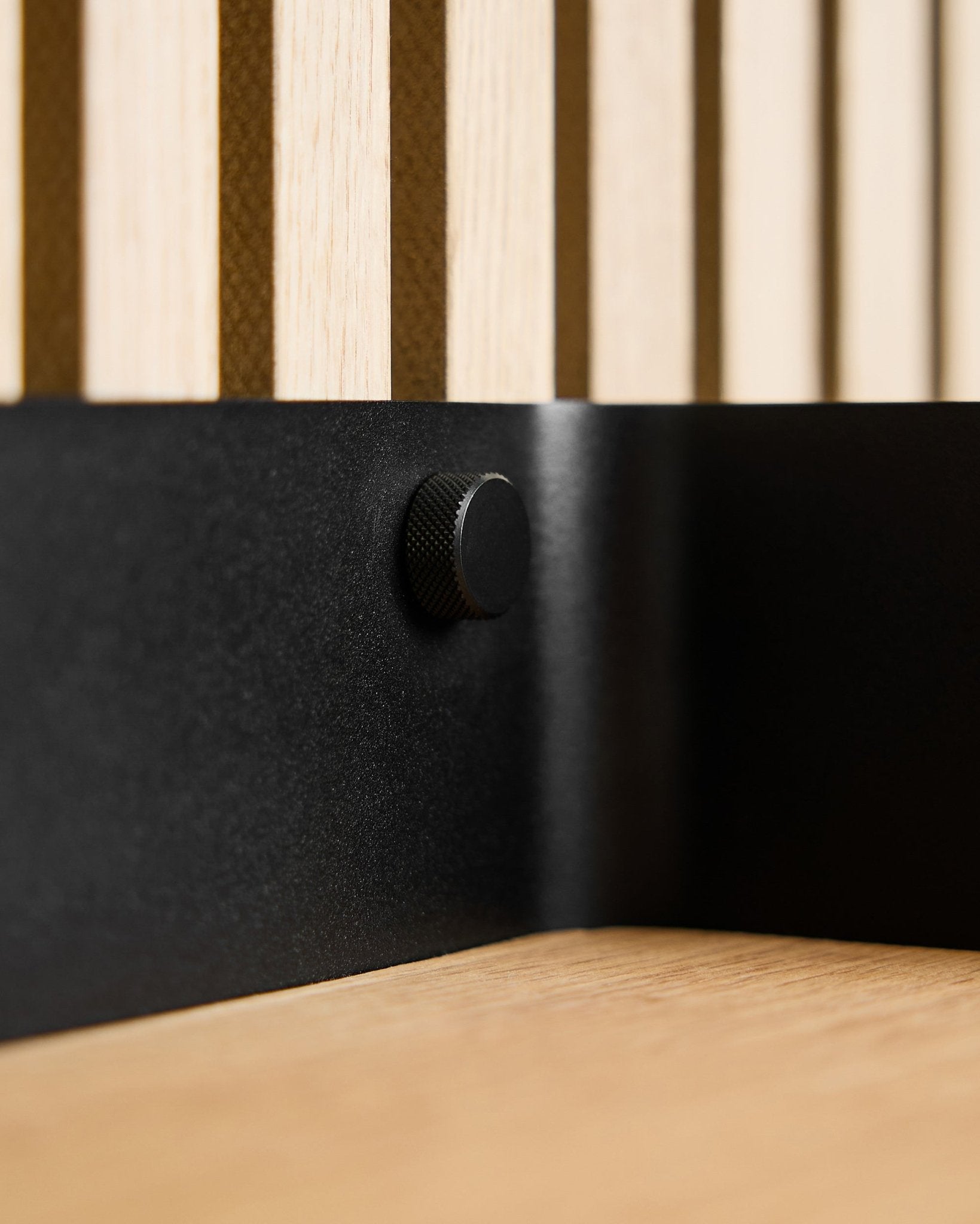 Nahaufnahme von zwei black WoodUpp Finger-Schrauben (M5x10 mm, 20x8,8) mit brass Schrauben, platziert auf einer nordischen Akustik-Lamellenwand aus Holz und einer hellen Holzoberfläche.