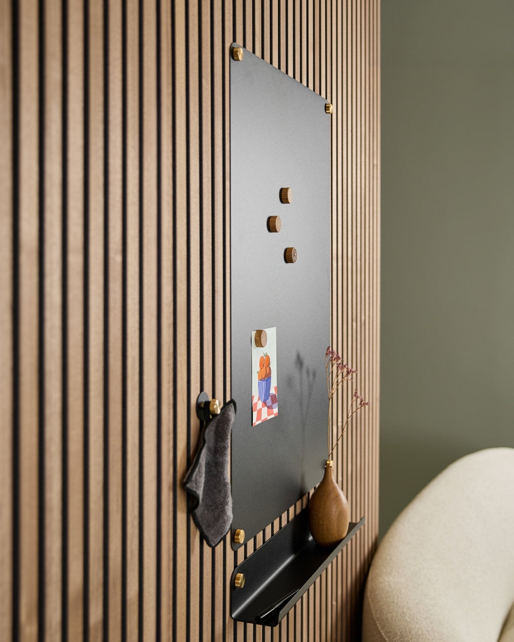 Die Norr large black akustische Magnettafel von WoodUpp mit Holzmagneten, Pinnwand, Regal und Vase ist neben einem beige Stuhl an der Wand montiert.