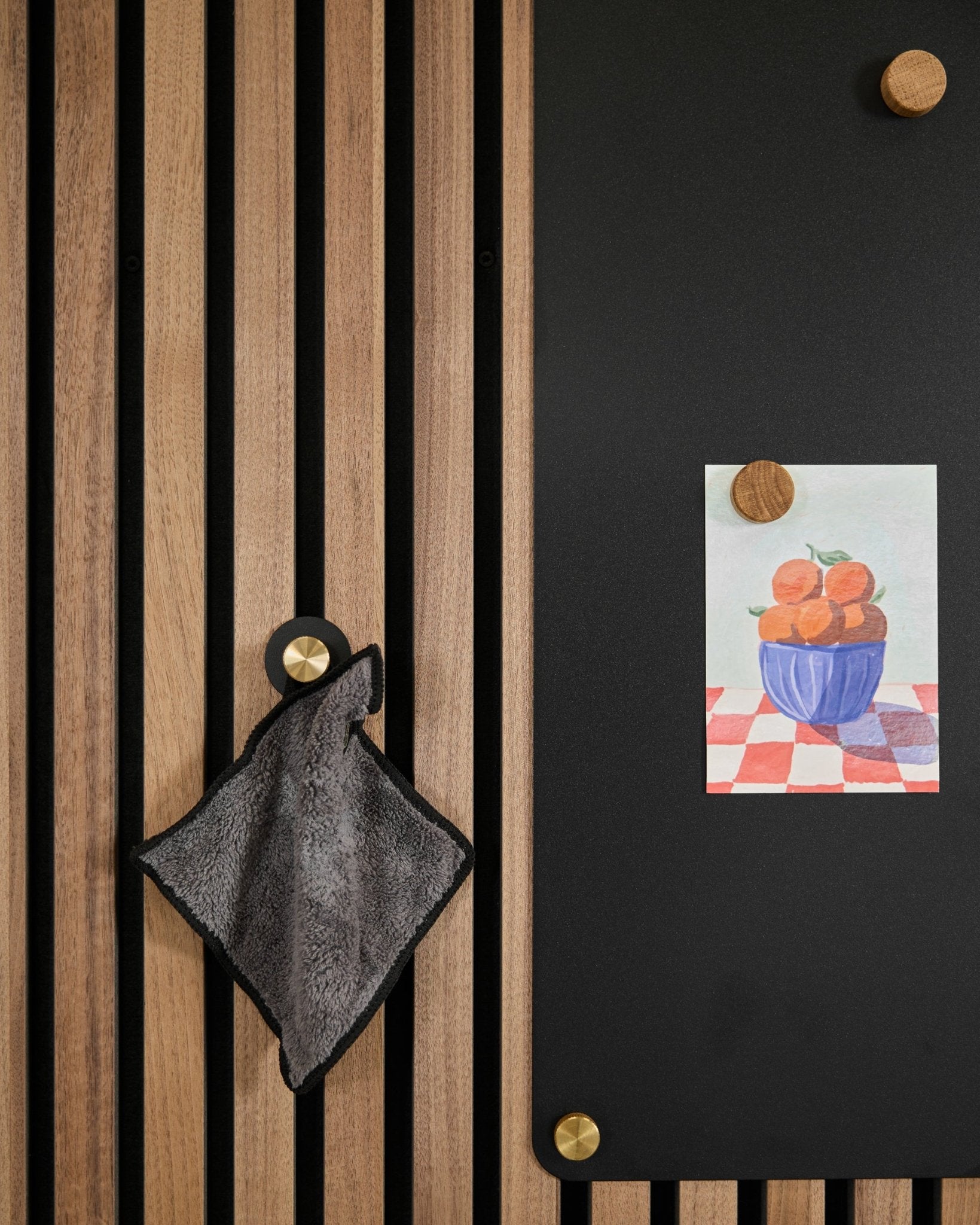 Ein small Handtuch hängt an einem nordischen Akustik-Lamellenpaneel neben einer black Tafel, auf der eine Zeichnung von Orangen in einer Schale mit zwei goldenen Magneten befestigt ist.
