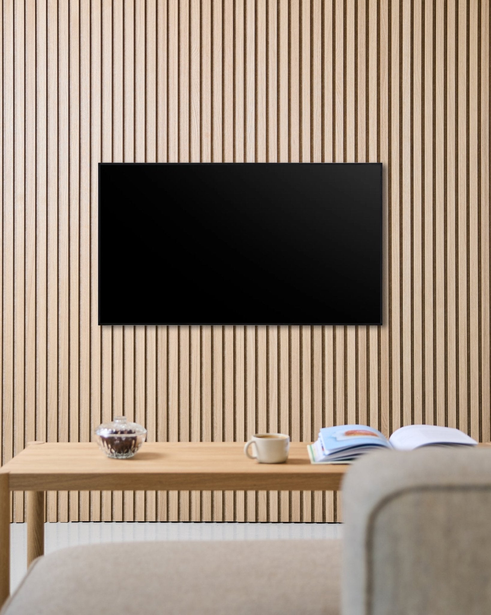 Eine Fjell TV-Halterung (small) von WoodUpp befestigt einen Flachbildfernseher an einer nordischen Akupanel Akustikholzlamellenwand, über einem Tisch mit Tasse, Schale und offenem Magazin neben einem beige Sofa.