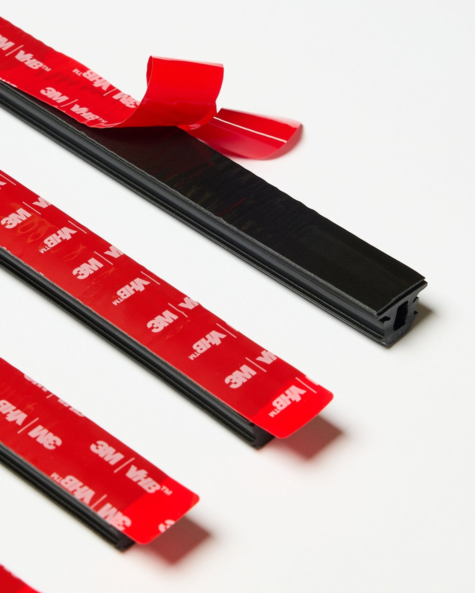 Set aus vier black WoodUpp CREATE Pop-on Profile Leisten mit sichtbarer roter 3M-Klebefläche, eine Leiste teilweise abgezogen, auf einer weißen Oberfläche präsentiert.