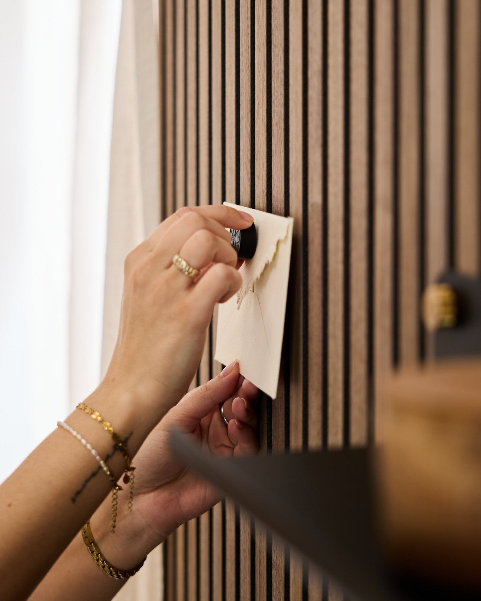 Eine Person mit goldenen Armbändern und Ringen befestigt einen Umschlag mit dem Norr Poster Magnet-Set in Black Oak an einem Eichen-Akustikpaneel mit Holzlamellen von WoodUpp.