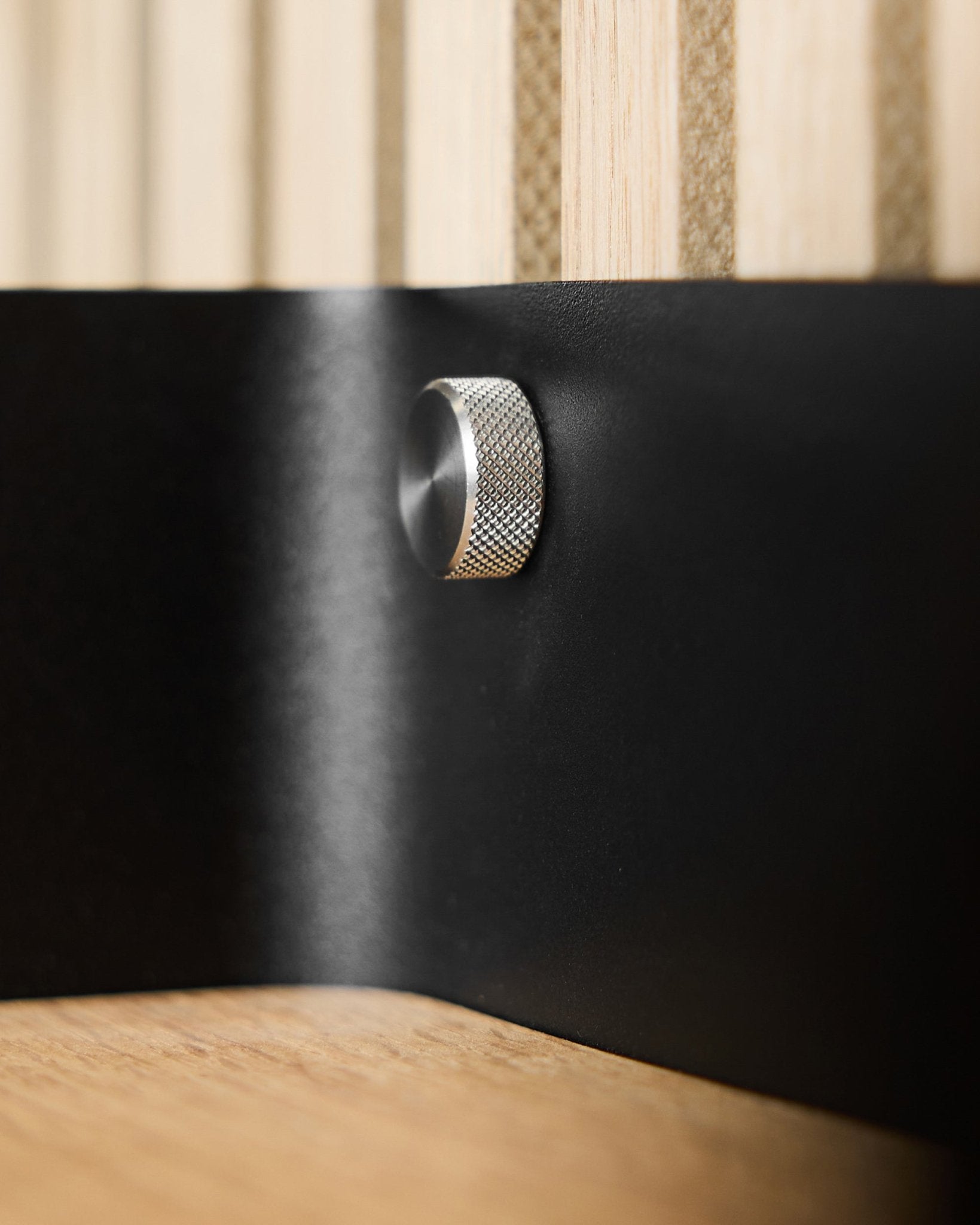 Nahaufnahme von zwei WoodUpp Fingerschrauben (M5x10 mm, 20x8.8 Edelstahl steel) auf einer black Metalloberfläche, mit Akupanel akustischen Holzlamellen im Hintergrund.