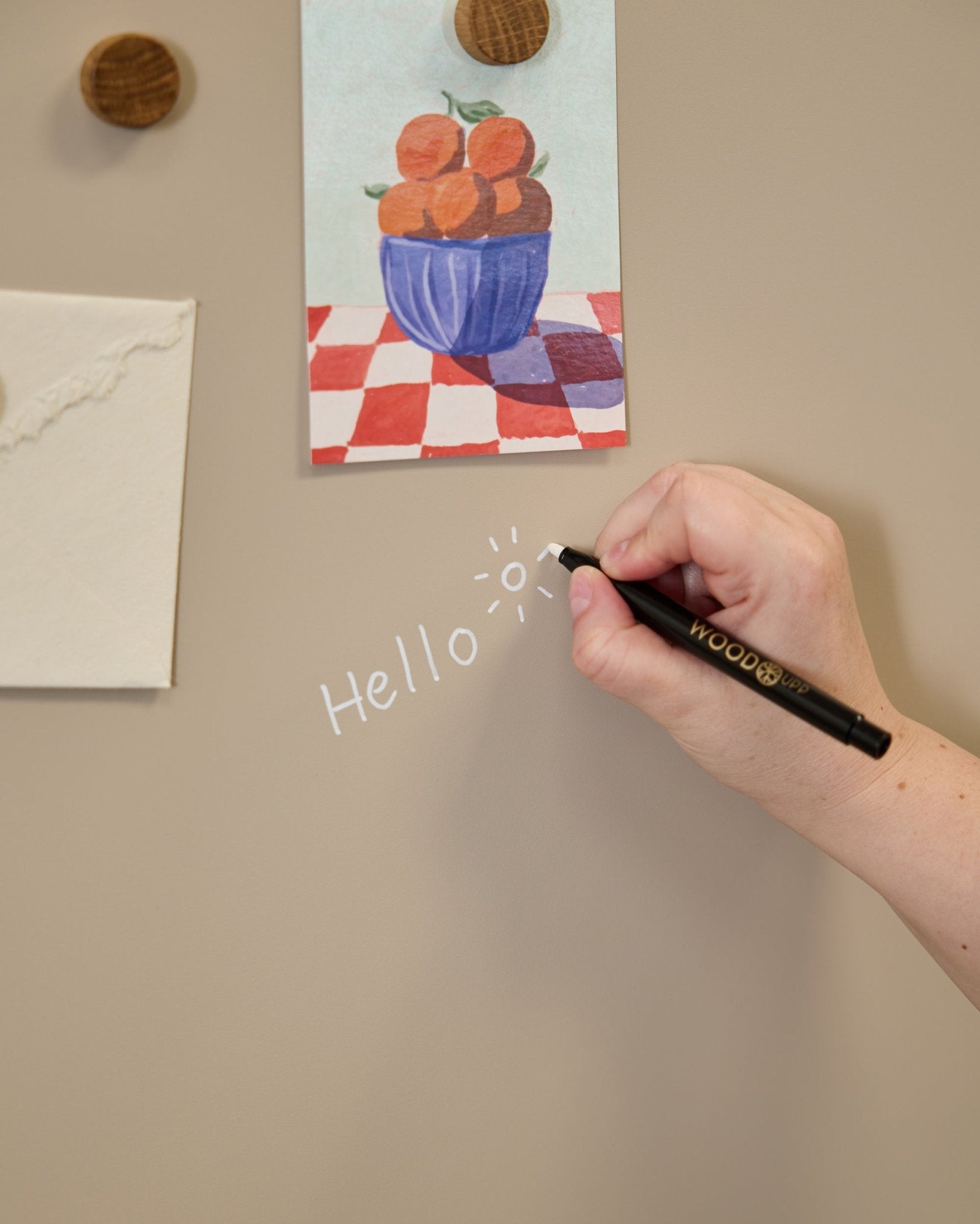 Eine Hand schreibt hello mit einer Sonnenkritzelei auf das WoodUpp Norr Blackboard, daneben Kunstwerke, die mit runden Magneten an einem Akustikpaneel befestigt sind.