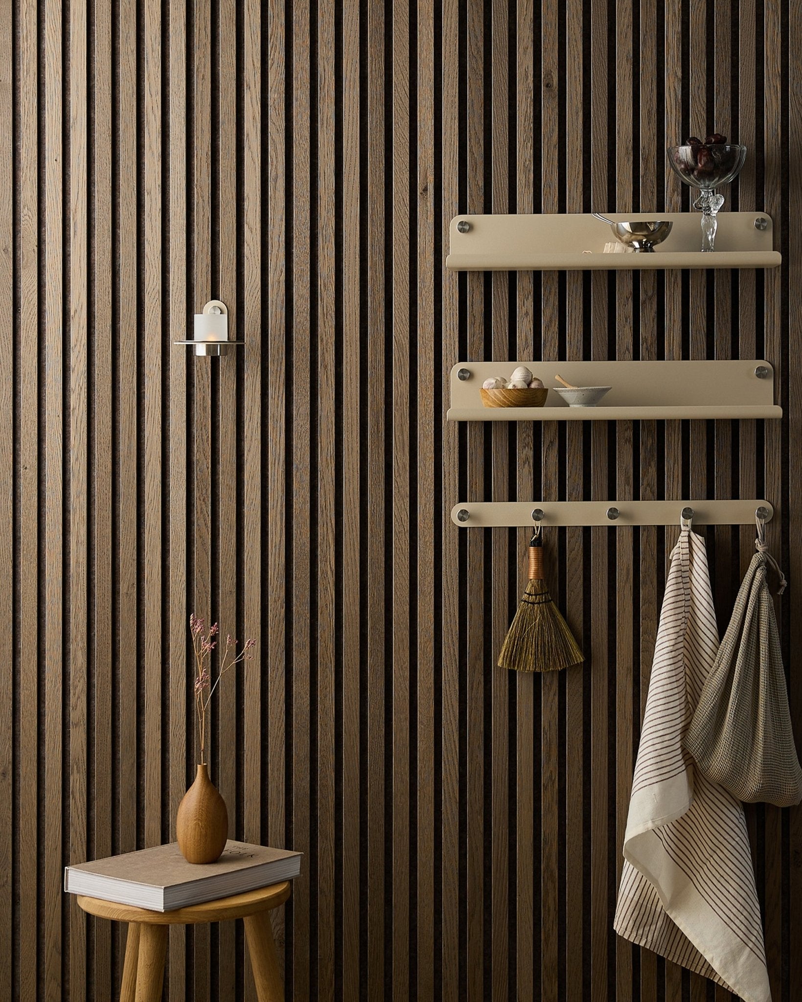 Ein Holzhocker mit Trockenblumen steht neben einem WoodUpp Akupanel Akustikpaneel, neben beige Regalböden mit Geschirr, einer Bürste, Handtüchern und einem Norr Kerzenhalter.