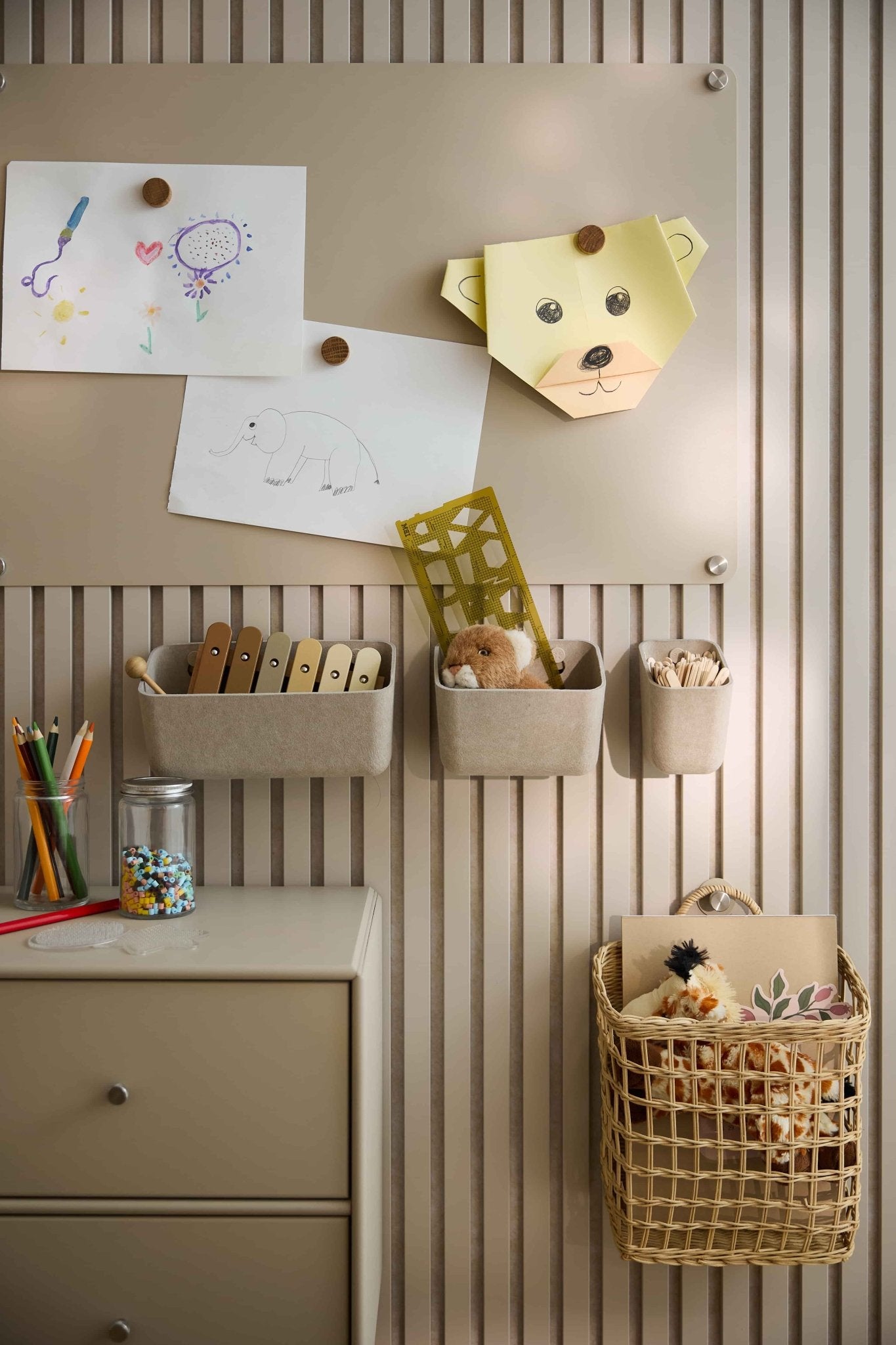 An einer beigen Wand mit einer WoodUpp-Akustikplatte sind Kinderkunstwerke und Spielzeug angeordnet, darunter Filzbehälter, Körbe, eine Schublade und eine Lochwand.