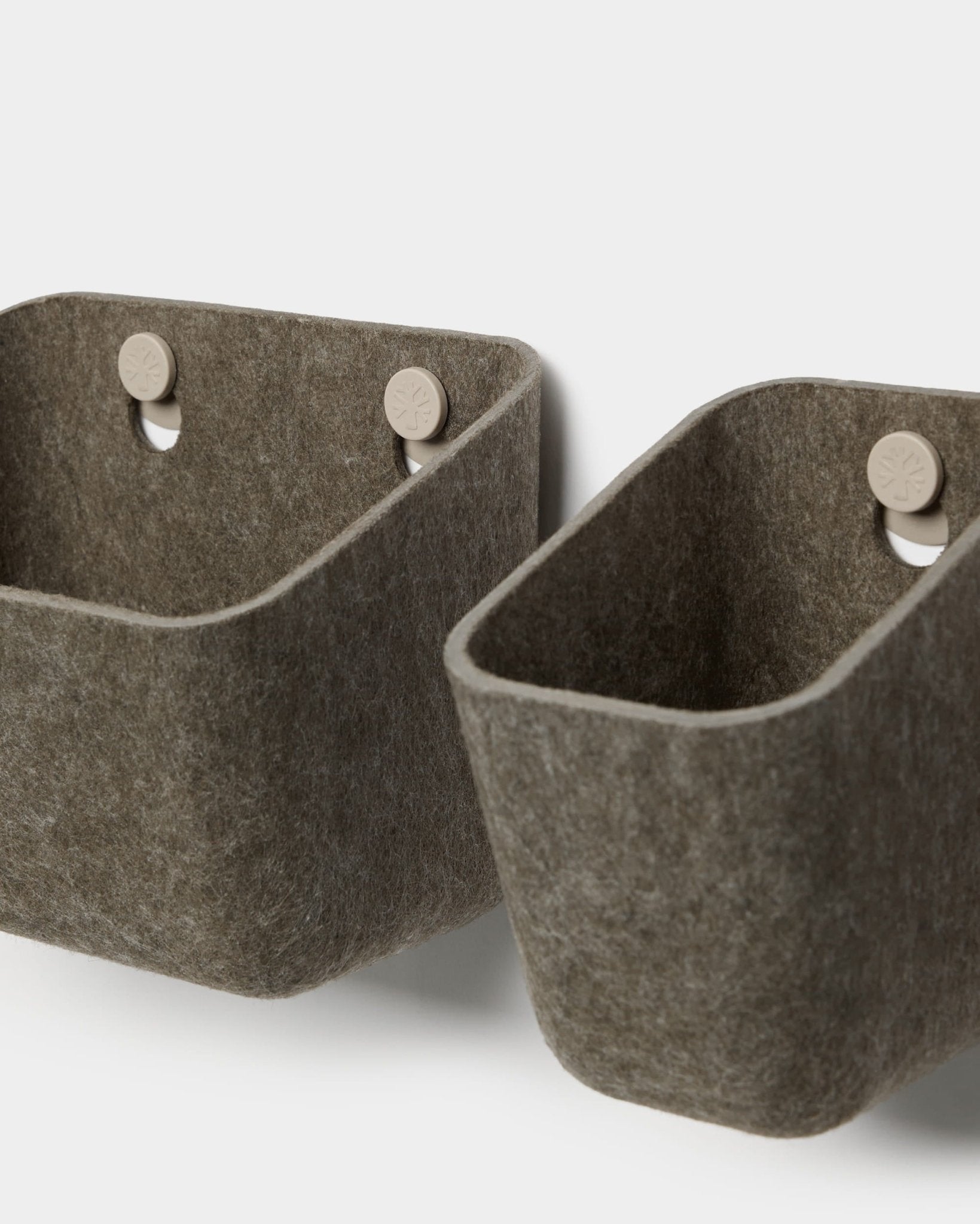 Zwei Luva-Filzbehälter in dunklem, warmem Grau von WoodUpp hängen an beigen Haken, die an einer Akustikplatte befestigt sind, und bieten so eine minimalistische Wandablage mit abgerundeten Ecken.