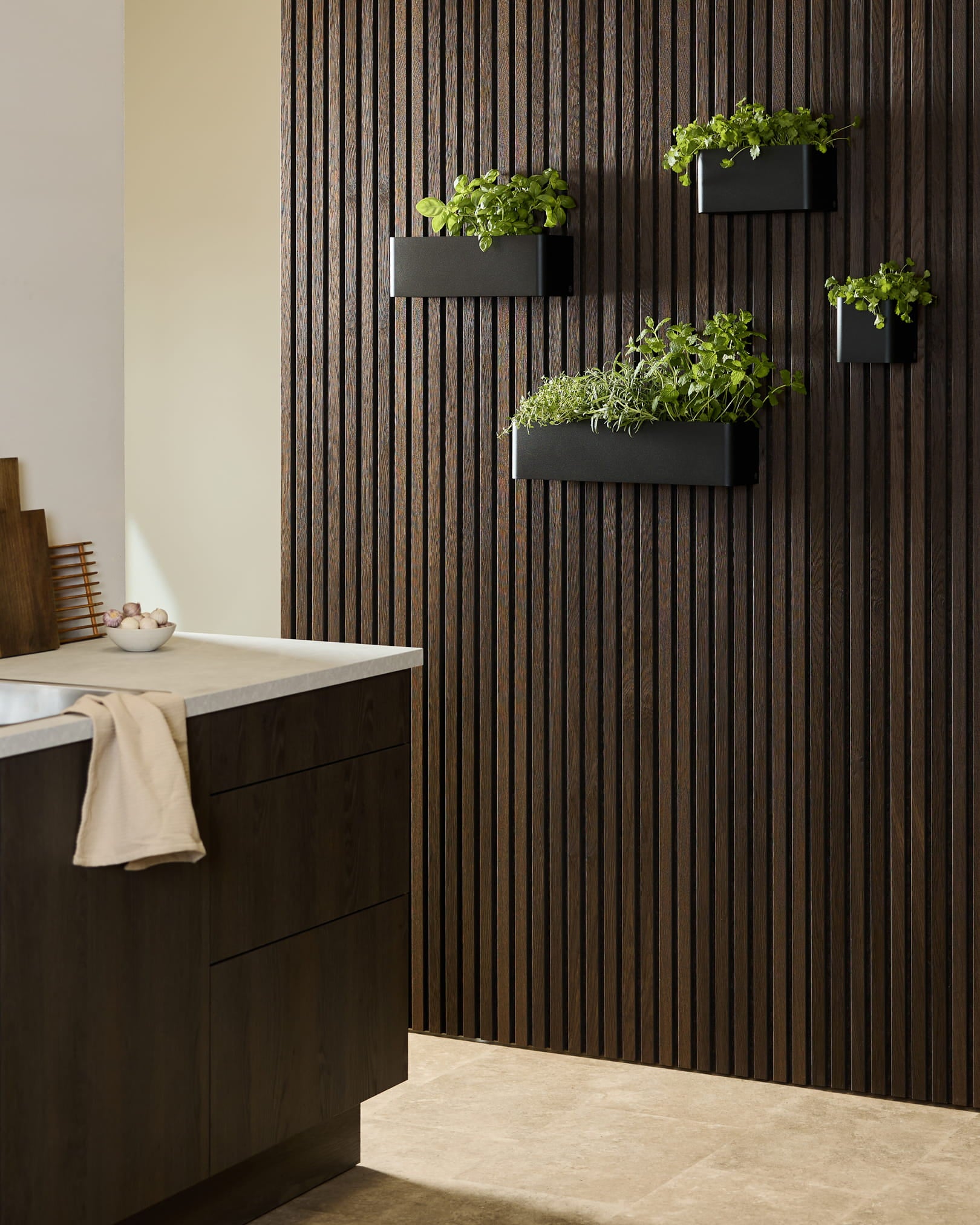 Moderne Küche mit einem dunklen WoodUpp Akupanel Holz-Akustikpaneel, einer black Norr Plant Box mit Kräutern und dunklen Schränken mit weißer Arbeitsplatte.