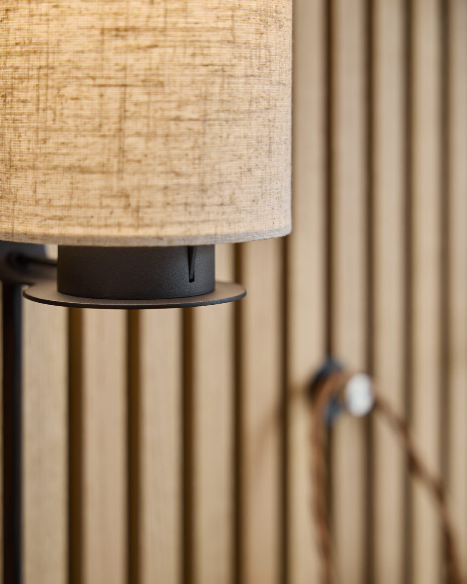 Feya Wandleuchte / Black/Beige von WoodUpp mit einem Leinenschirm aus Baumwollmischung in beige, einem Metallsockel in black und sichtbarem Kabel, montiert auf einer nordischen Akustik-Lamellenwand.