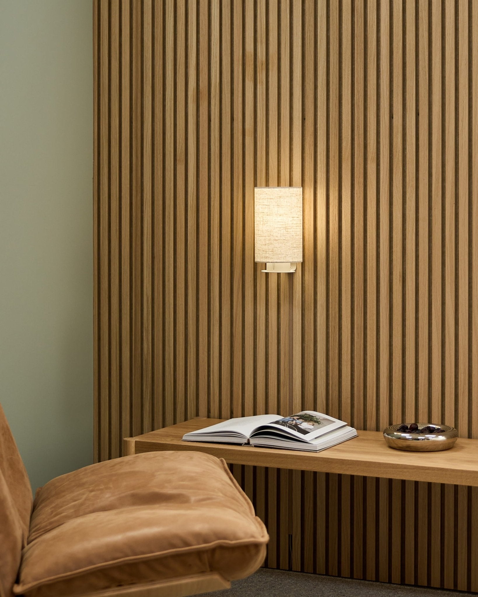 Minimalistischer nordischer Raum mit einem hellbraunen Stuhl, einer Akupanel Akustik-Lamellenwand von WoodUpp, der Feya Wandleuchte in Beige/Beige, einem offenen Buch auf einer Bank und einer Schale.