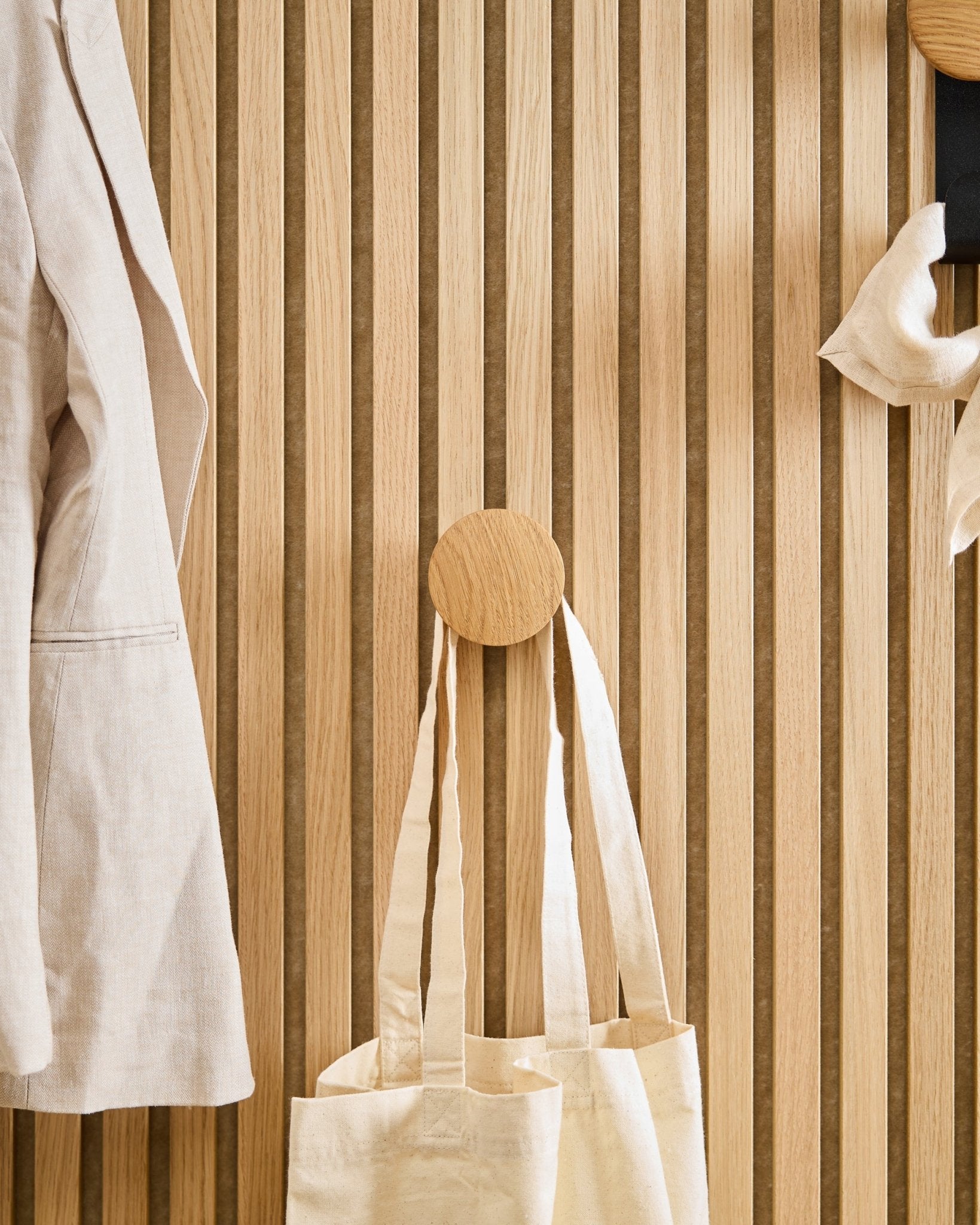 Ein beige Mantel und eine Tote Bag hängen an dem Linn I Single Hanger in Natural Oak von WoodUpp, montiert auf einem vertical Akupanel Akustikpaneel mit Holzlamellen.