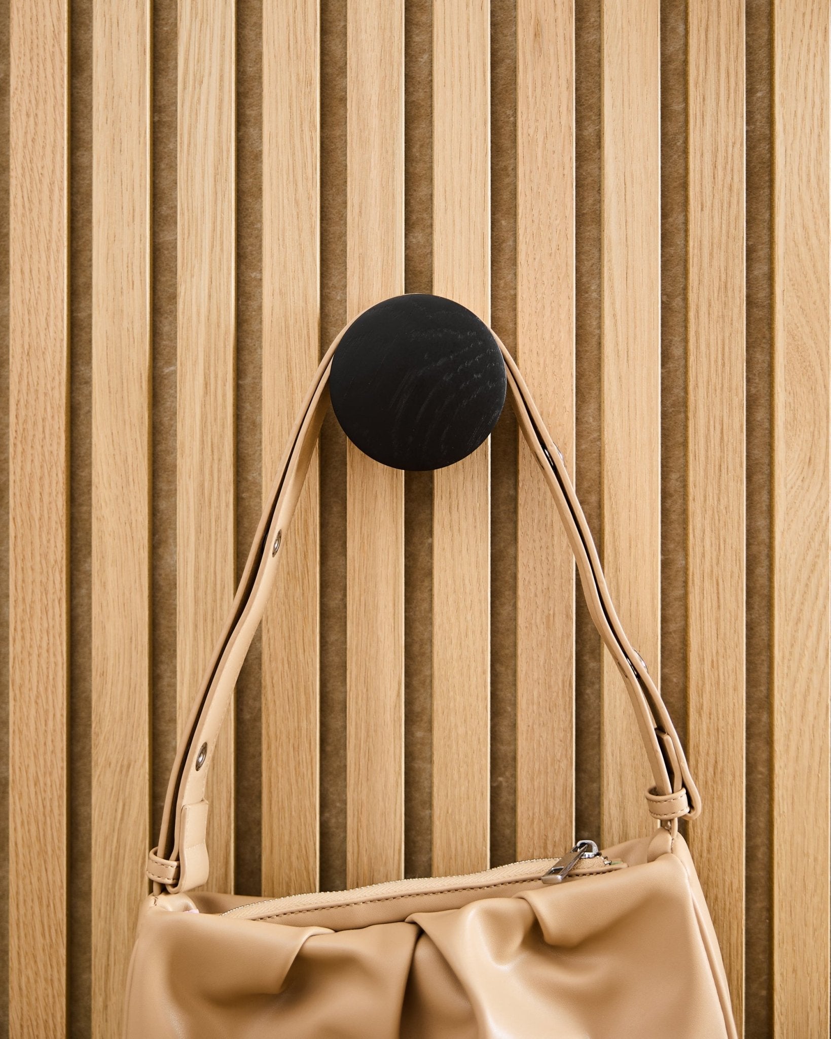 Beige Handtasche hängt an einem WoodUpp Linn | Single Hanger in Black Oak, montiert auf einem Akustikpaneel mit Holzlamellen in hellem Holzton Akupanel.