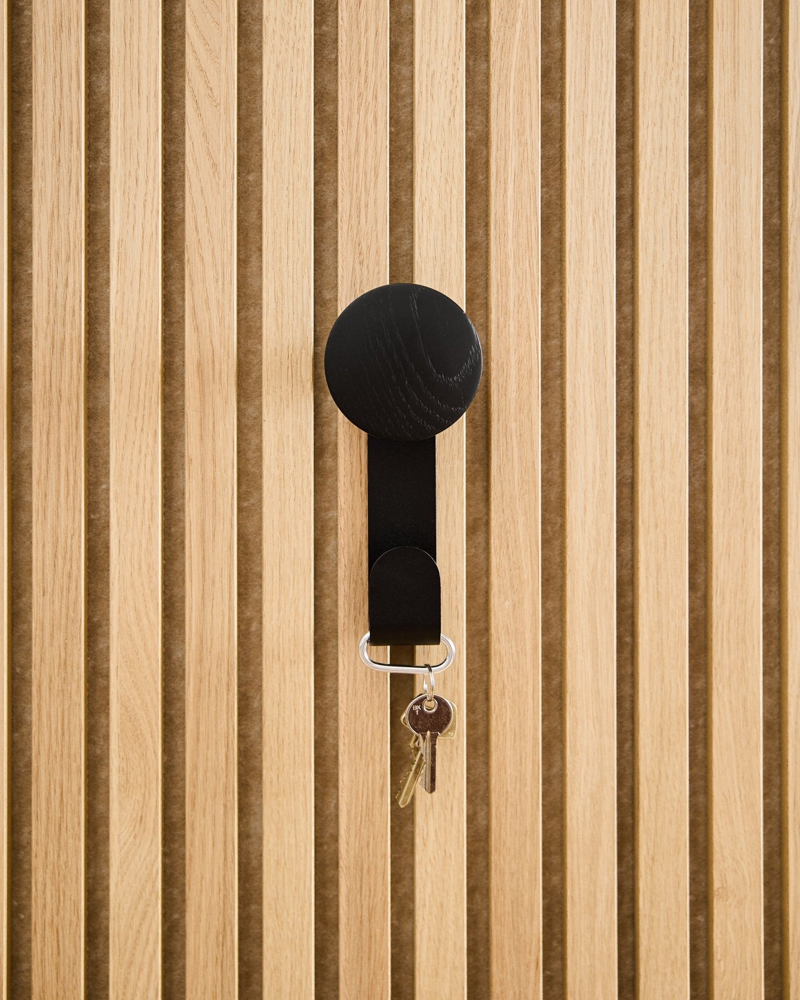 Ein Linn Vertical Haken in Black Oak von WoodUpp trägt einen Schlüsselbund mit zwei Schlüsseln und einem runden Anhänger und ist an einem strukturierten Akupanel Akustikholzpaneel montiert.