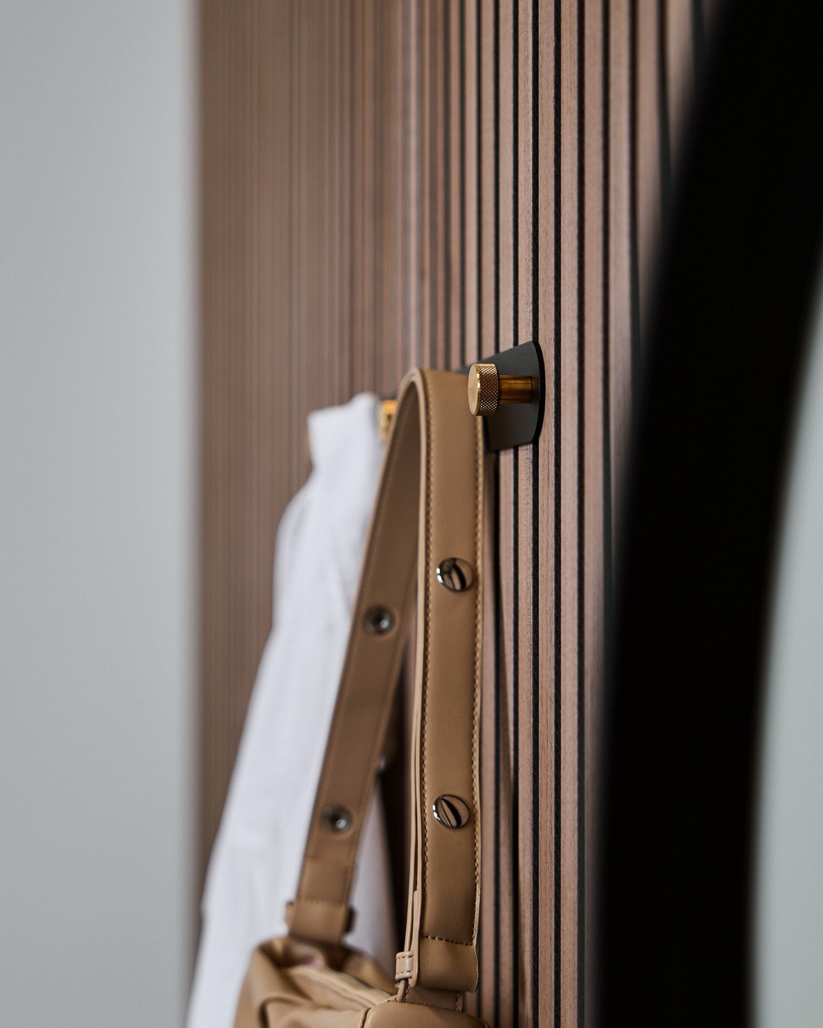 Eine hellbraune Tasche und ein weißes Hemd hängen an einer WoodUpp Norr Garderobenleiste (Black/Brass, X5), die an einem vertical Akupanel Akustikpaneel mit Holzlamellen montiert ist.
