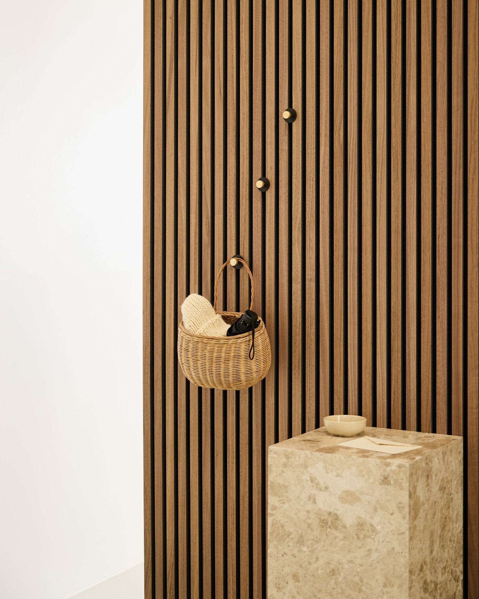 Norr Kleiderhaken von WoodUpp, montiert an einer nordischen Akupanel Akustik-Lamellenwand, neben einem beige Steinsockel mit einer small Schale in einem minimalistischen Interieur.