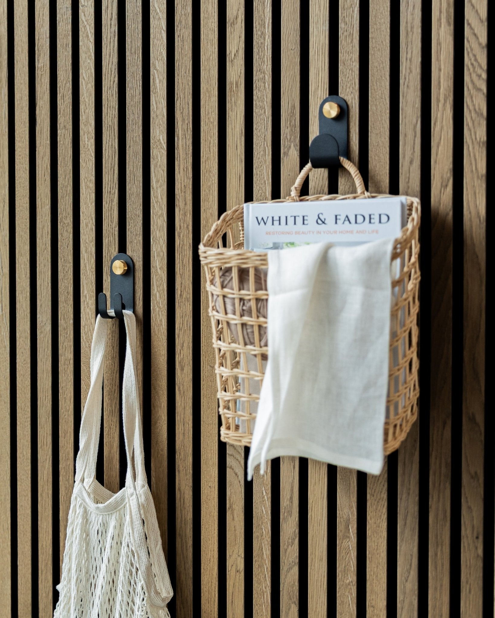 WoodUpps Norr Single Hanger in Black trägt einen geflochtenen Korb und eine Netztasche und ist für geordneten Stauraum an einem nordischen Akustikpaneel mit Holzlamellen montiert.