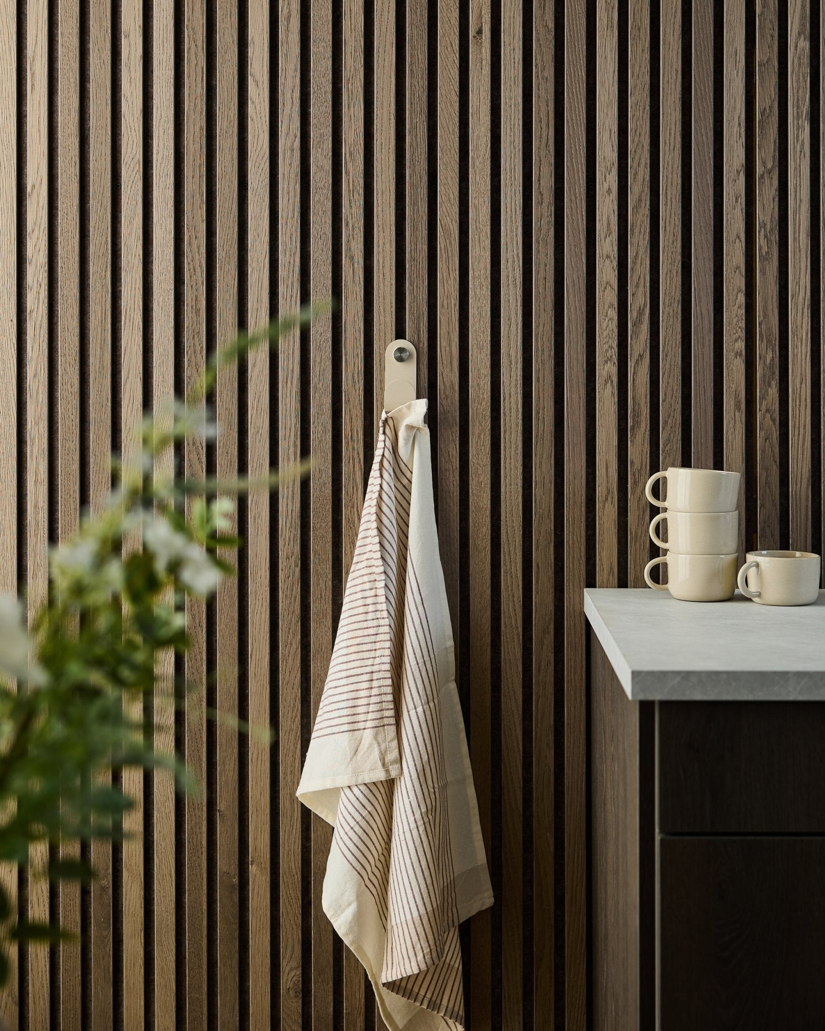 Ein gestreiftes Handtuch hängt an einem beige WoodUpp Norr Single Hanger, montiert an einem Holzlamellen-Akustikpaneel; darunter stehen gestapelte Tassen und eine Pflanze auf der Arbeitsplatte.