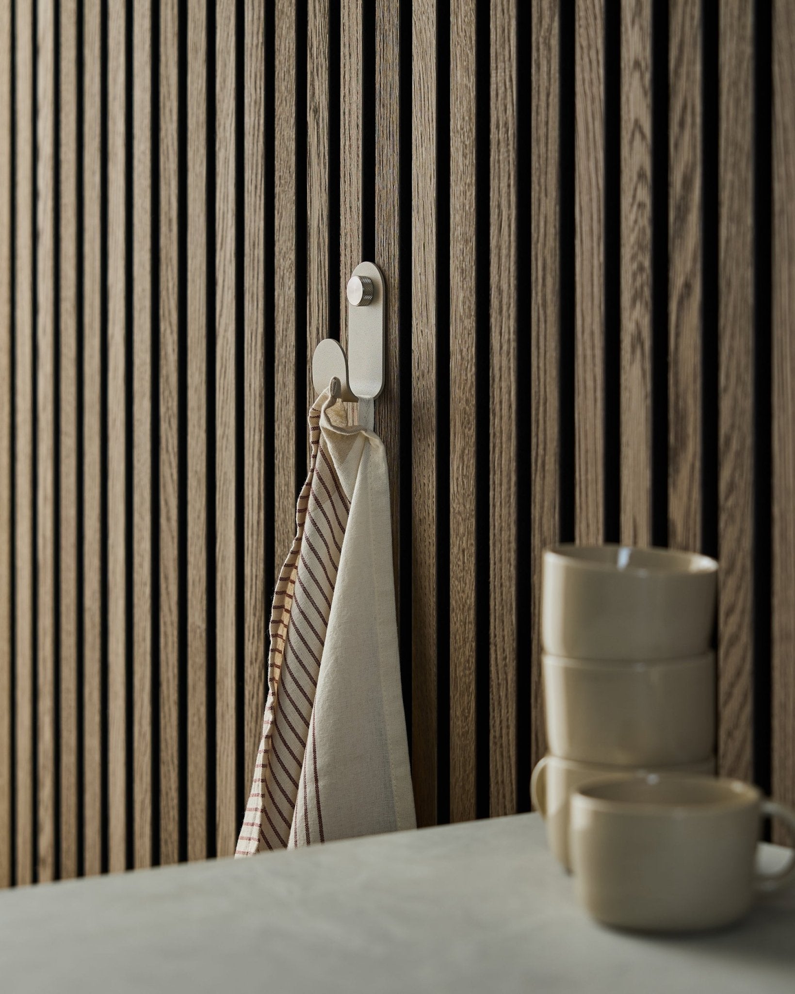 Ein gestreiftes Handtuch hängt an einem WoodUpp Norr Single Hanger in beige, montiert an einem Akustikpaneel im nordischen Lamellendesign; davor sind drei beige-Tassen gestapelt.