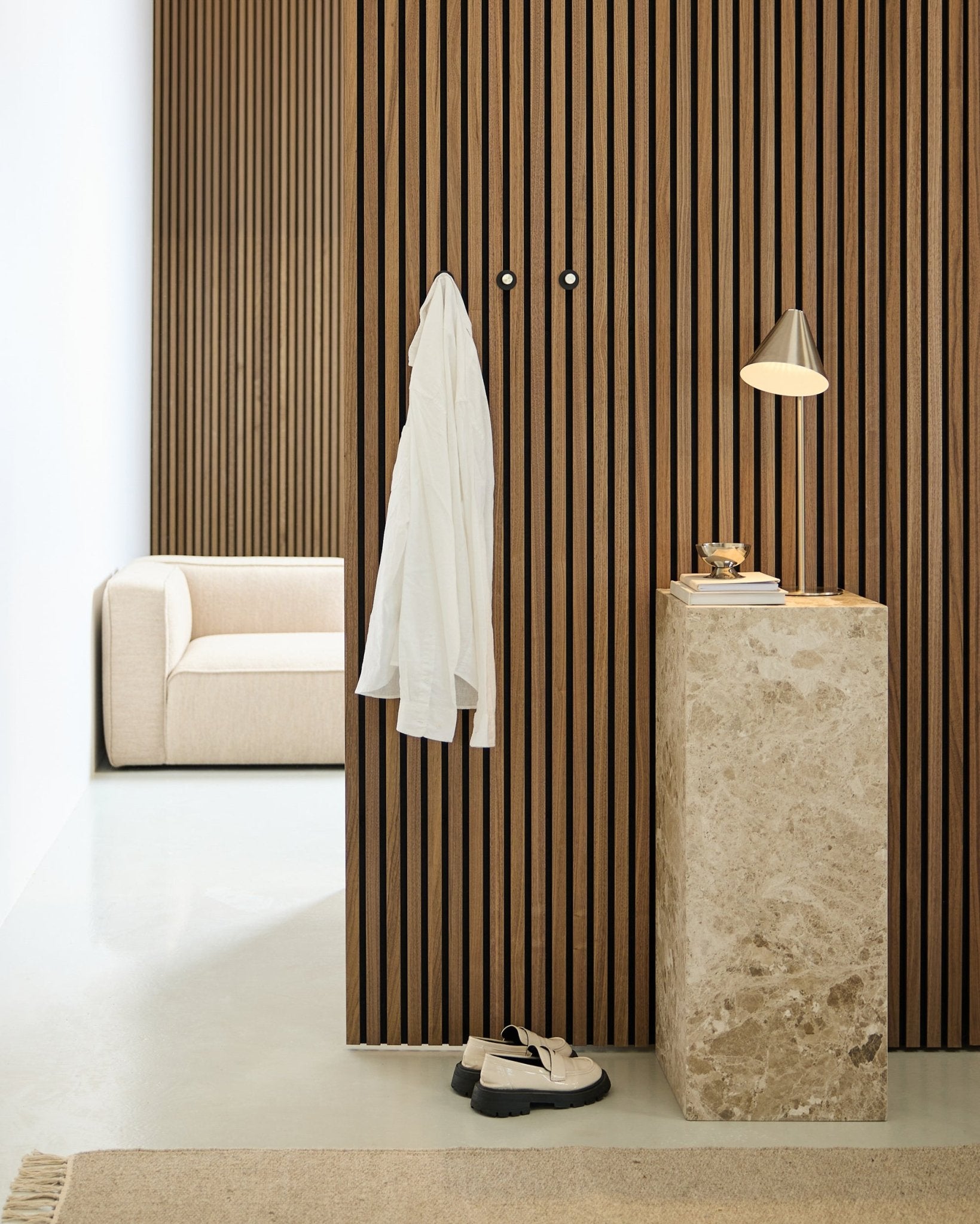 Minimalistischer Raum mit einer nordischen Akustikwand aus Holzlamellen in Akupanel, einem weißen Hemd an Wandhaken, einem Steinsockel mit Lampe und Büchern, einem Sofa in beige und weißen Schuhen auf dem Boden.