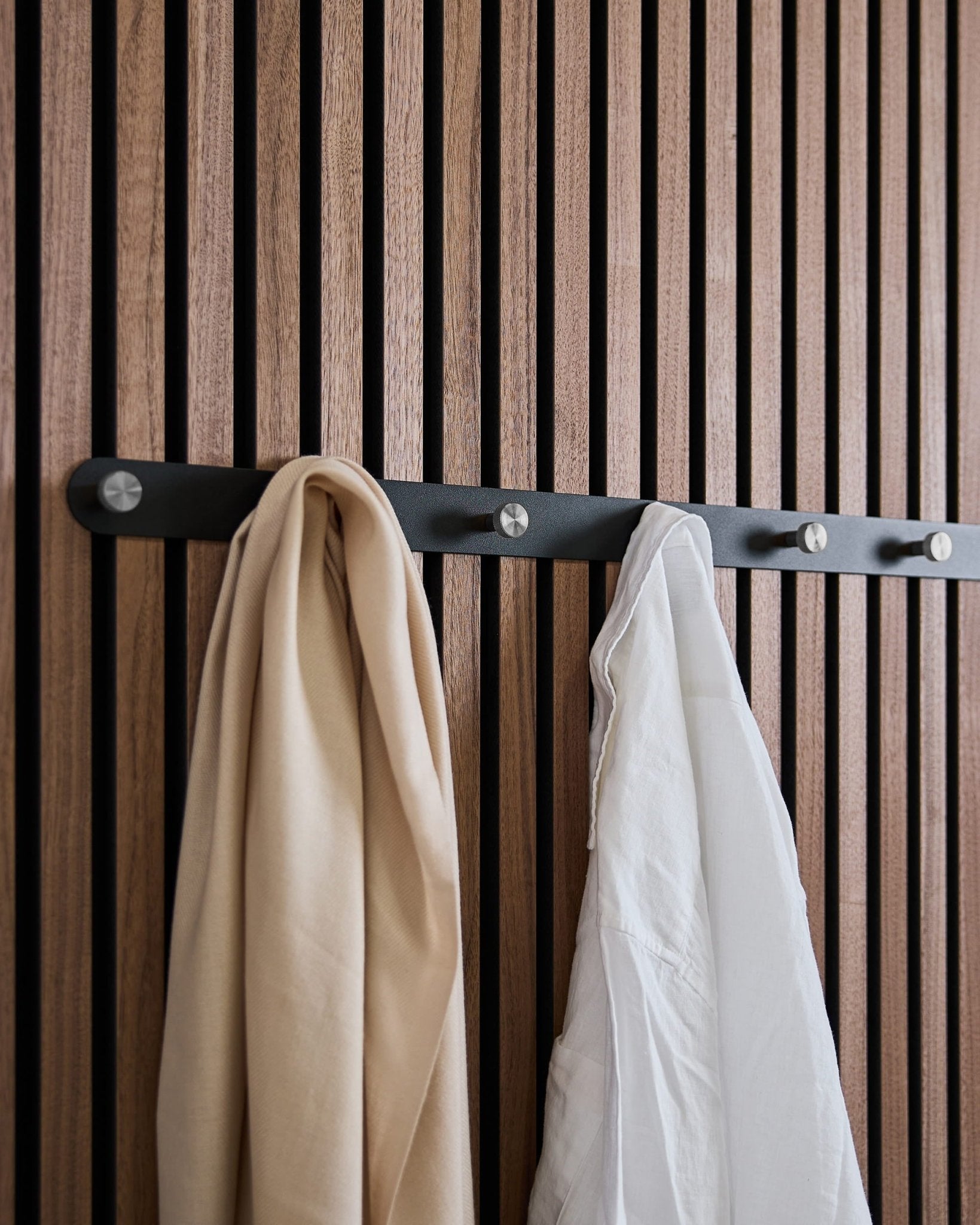 Ein beige Schal und ein weißes Hemd hängen an WoodUpps Norr X7 Black/Stainless Steel Garderobenleiste, die an einem nordischen Akustikpaneel mit Holzlamellen montiert ist.
