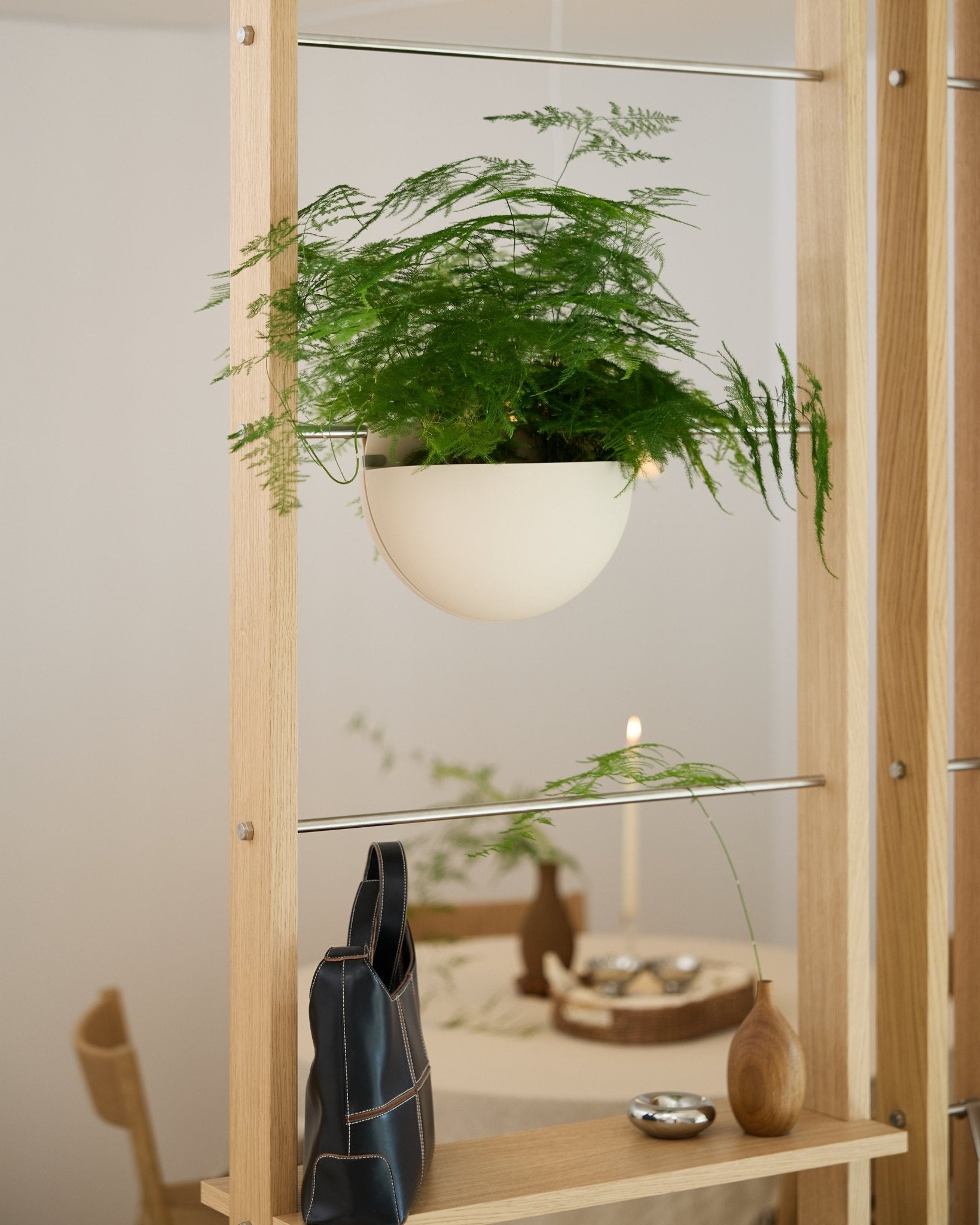 Ein grüner Farn in einem hellbeigen WoodUpp Gro Soft Pocket BM2-Pflanzgefäß steht auf einem Holzregal über einer schwarzen Tasche, einer Vase und einem gedeckten Esstisch.
