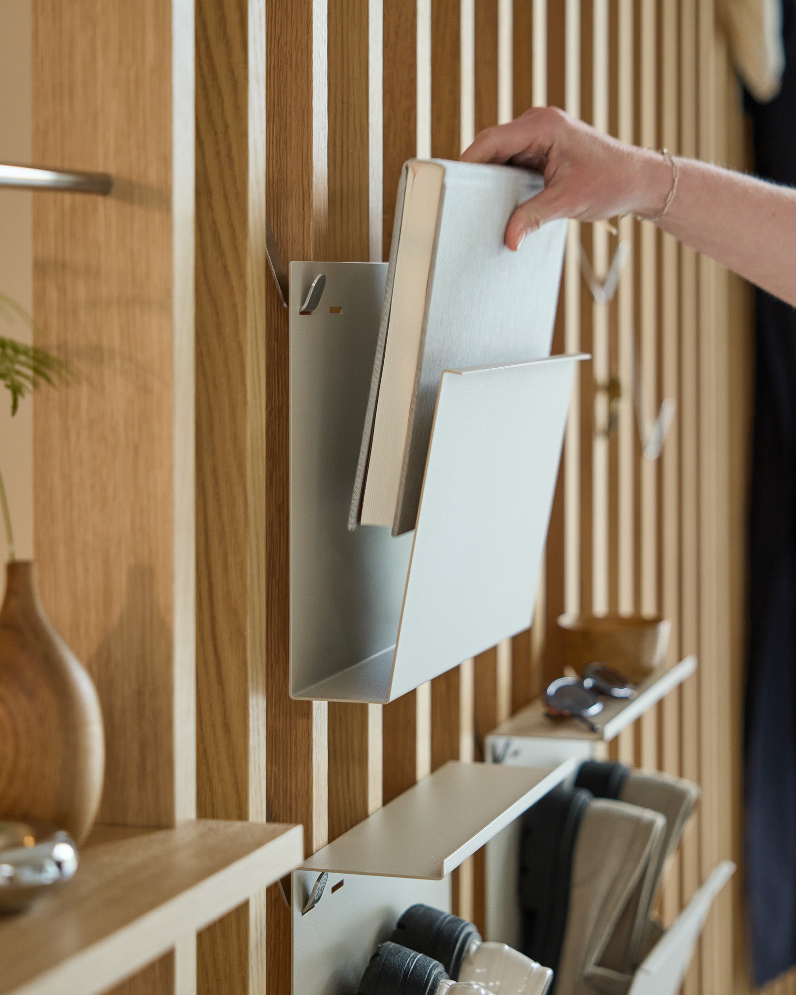 Eine Hand legt Nell | Magazine Storage / Beige von WoodUpp auf ein pulverbeschichtetes Regal aus steel, das an einem nordischen Holz-Akustikpaneel mit weiteren Gegenständen montiert ist.