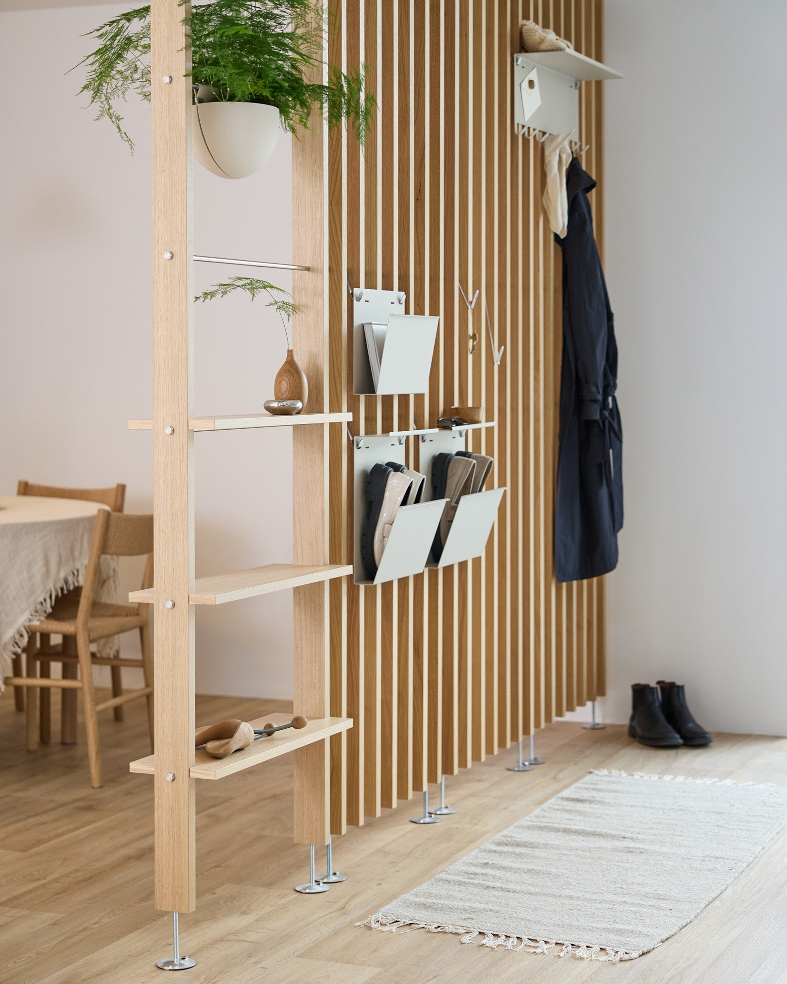 WoodUpp Nell Magazine Storage in beige, integriert in einen Raumteiler mit Regalböden aus steel, Aufbewahrungsboxen und Garderobenhaken; dahinter sind ein Essbereich und ein Teppich zu sehen.