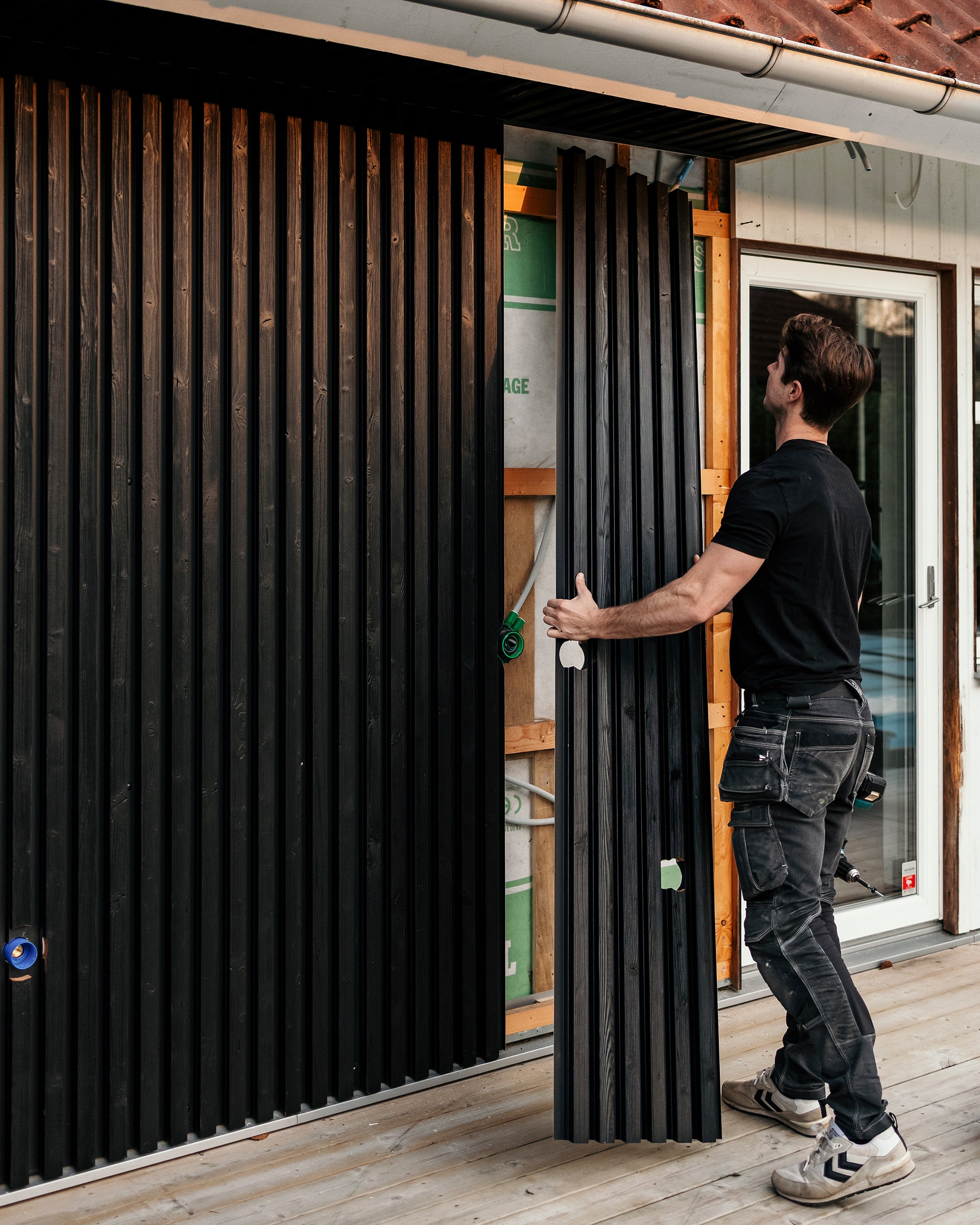 Ein Mann montiert vertikale schwarze Aluwood-Fassadenpaneele an der Außenwand eines Hauses, während er auf einem neu gebauten Deck steht.