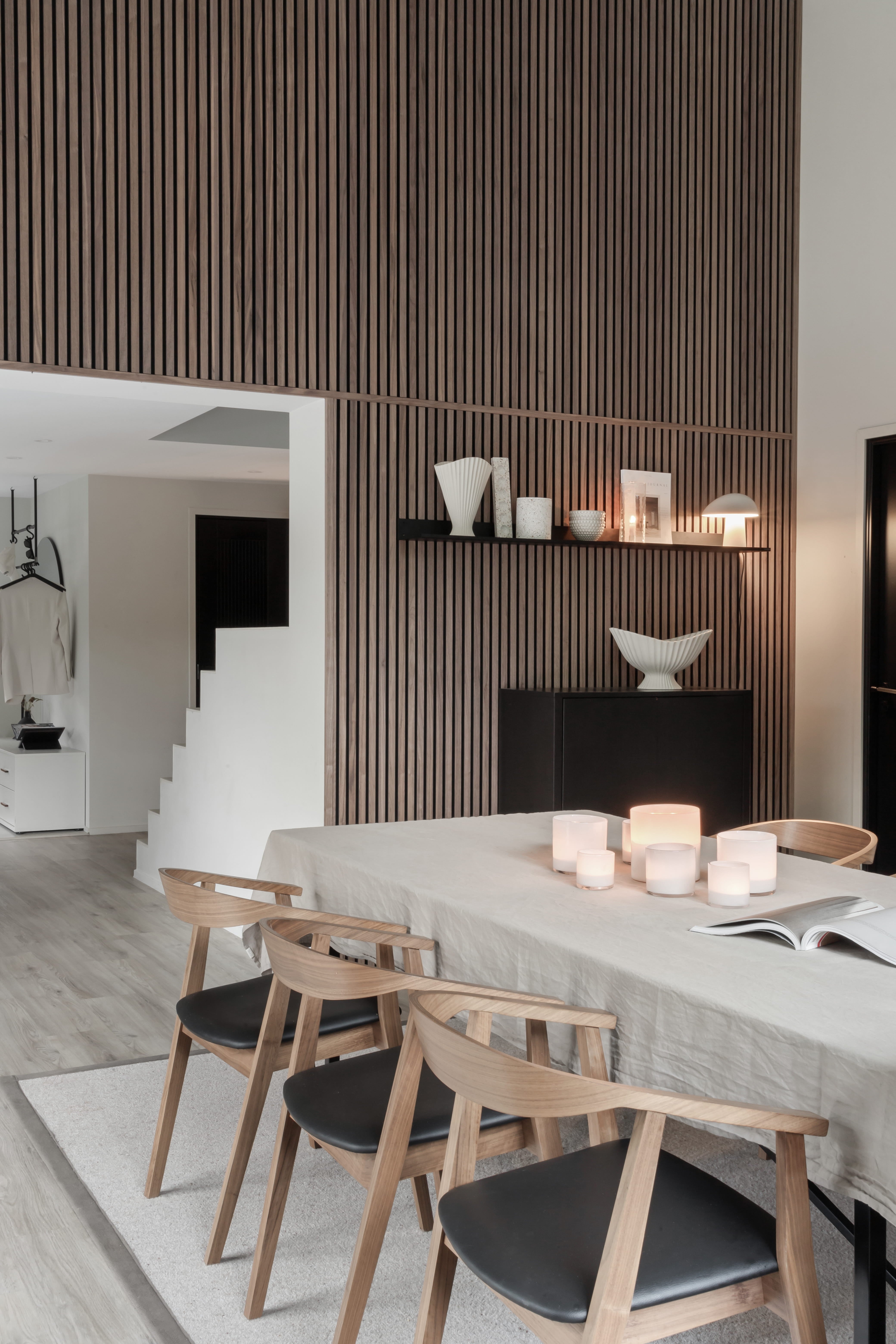 Modernes Esszimmer mit einem hellen Holztisch, vier Stühlen, Kerzen und minimalistischem Dekor. Vertikale Akupanel-Akustikholzlattenwand und offene Treppe im Blick.