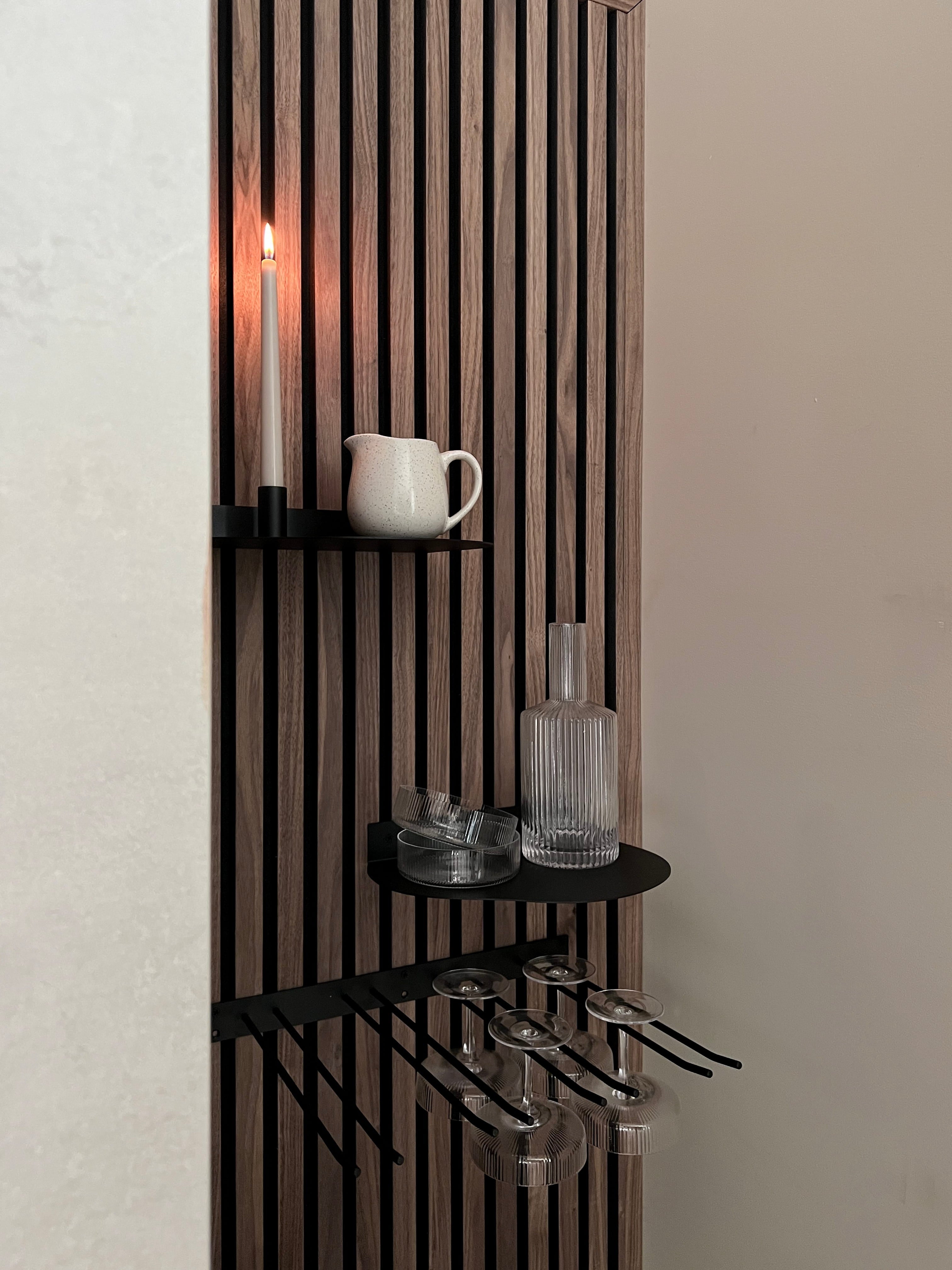 Modernes nordisches Akustikpaneel mit schwarzen Regalen, unter denen eine brennende Kerze, ein weißer Krug, eine Glasflasche und gestielte Weingläser hängen.